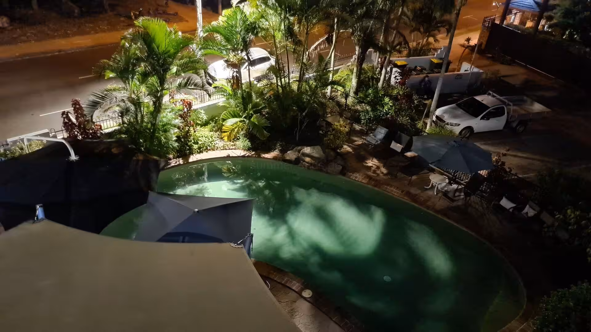 A lit turquoise pool glows among palms beside a street at night.