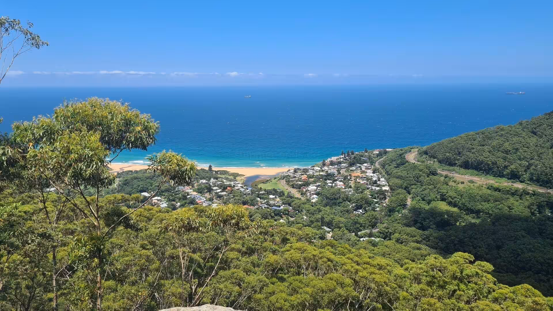 A sweeping view down to a turquoise bay, a sandy beach, and a small town ringed by forested green hills.