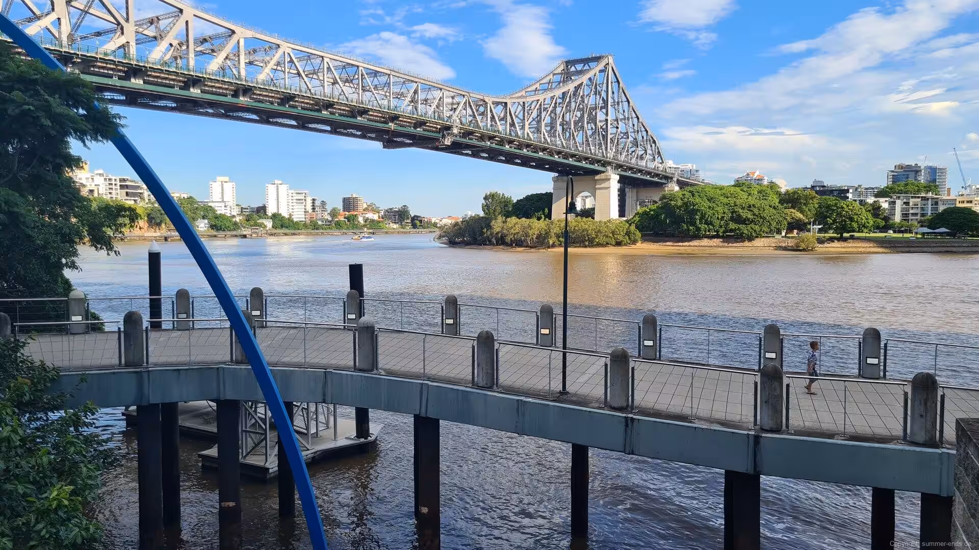 A steel truss bridge arches over a wide muddy river and a curved riverwalk.