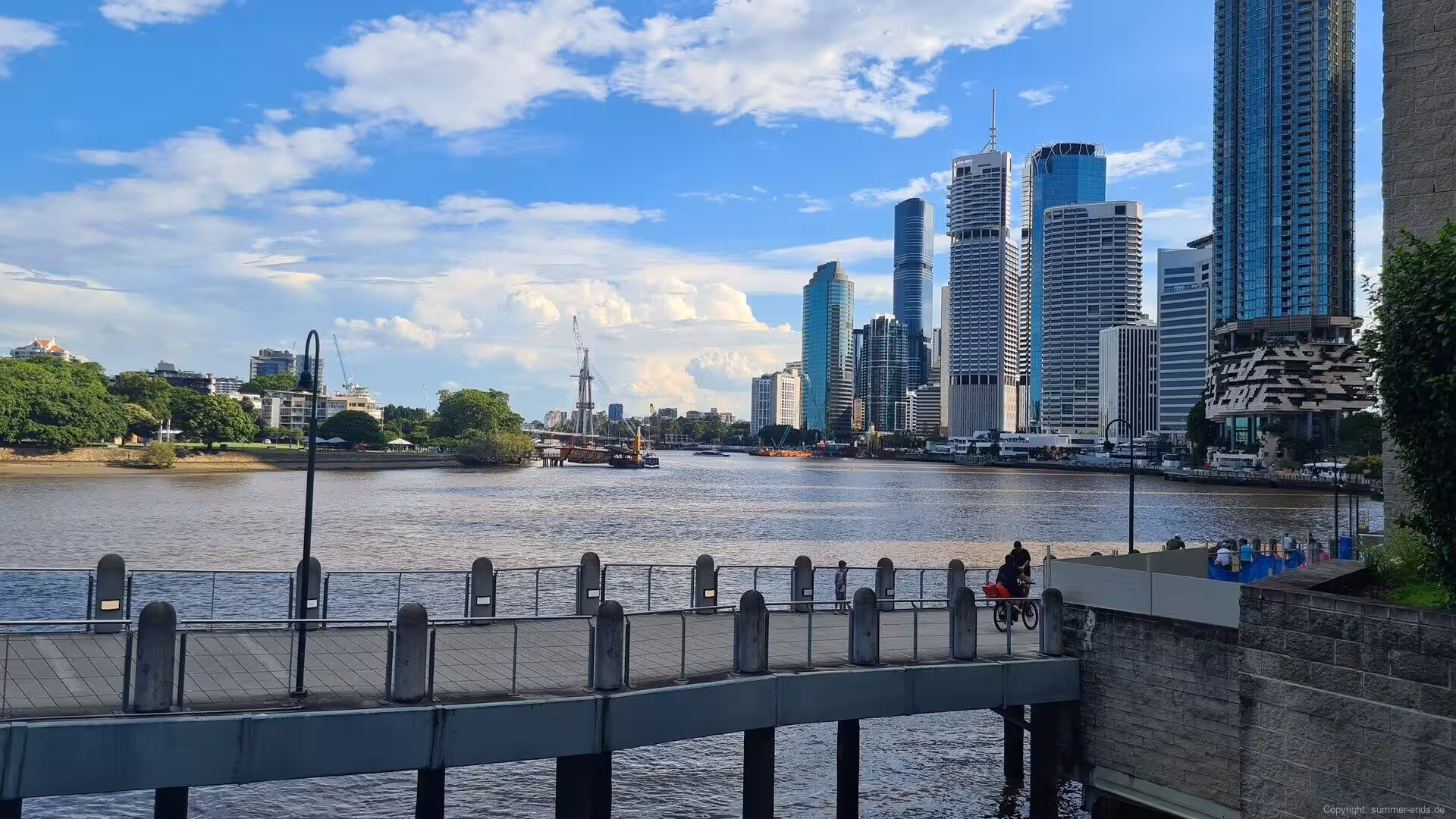 A curved riverside walkway fronts a skyline of glass towers across the water.
