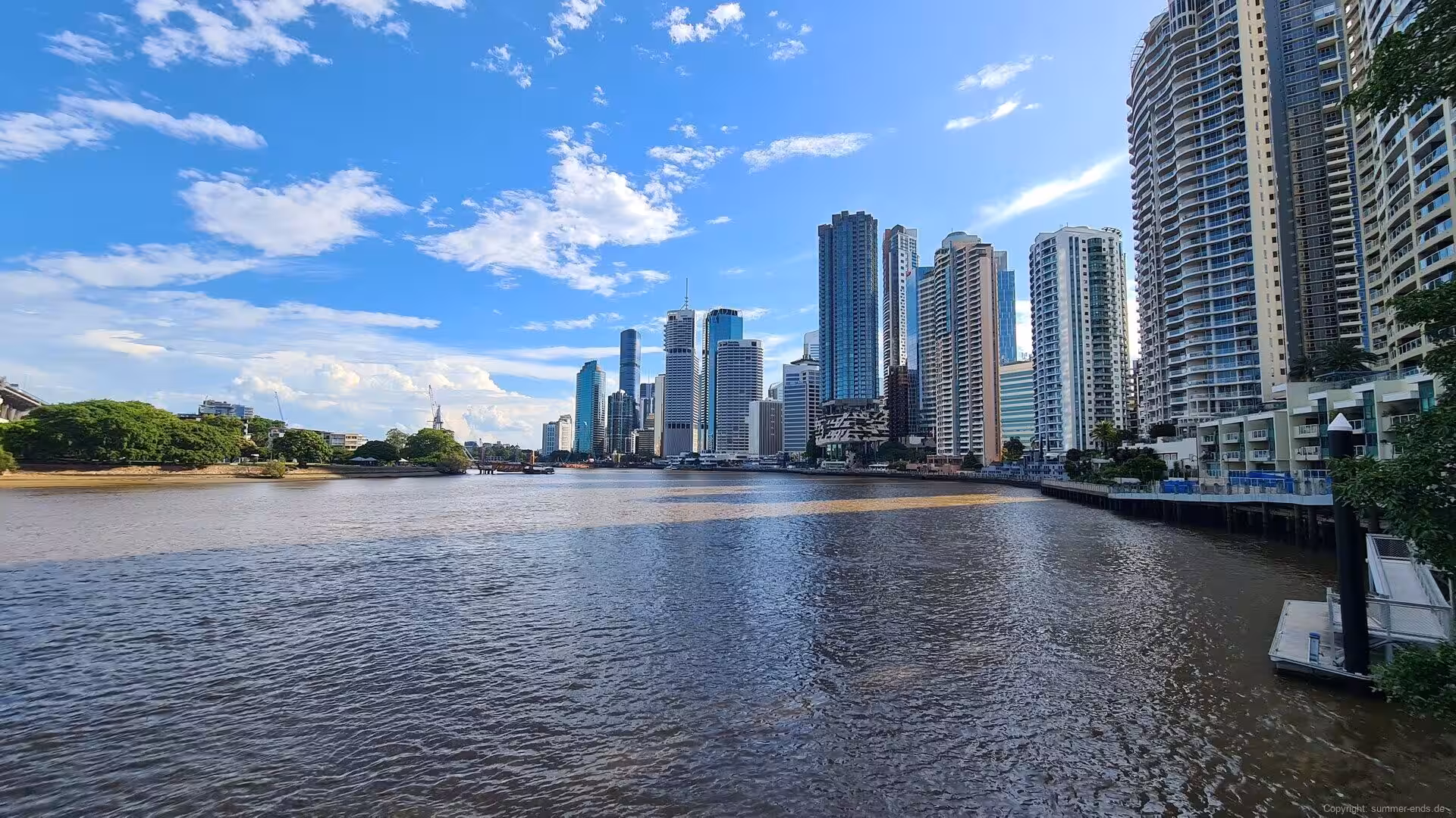 A dense line of glass towers rises along a broad river beneath a blue sky.