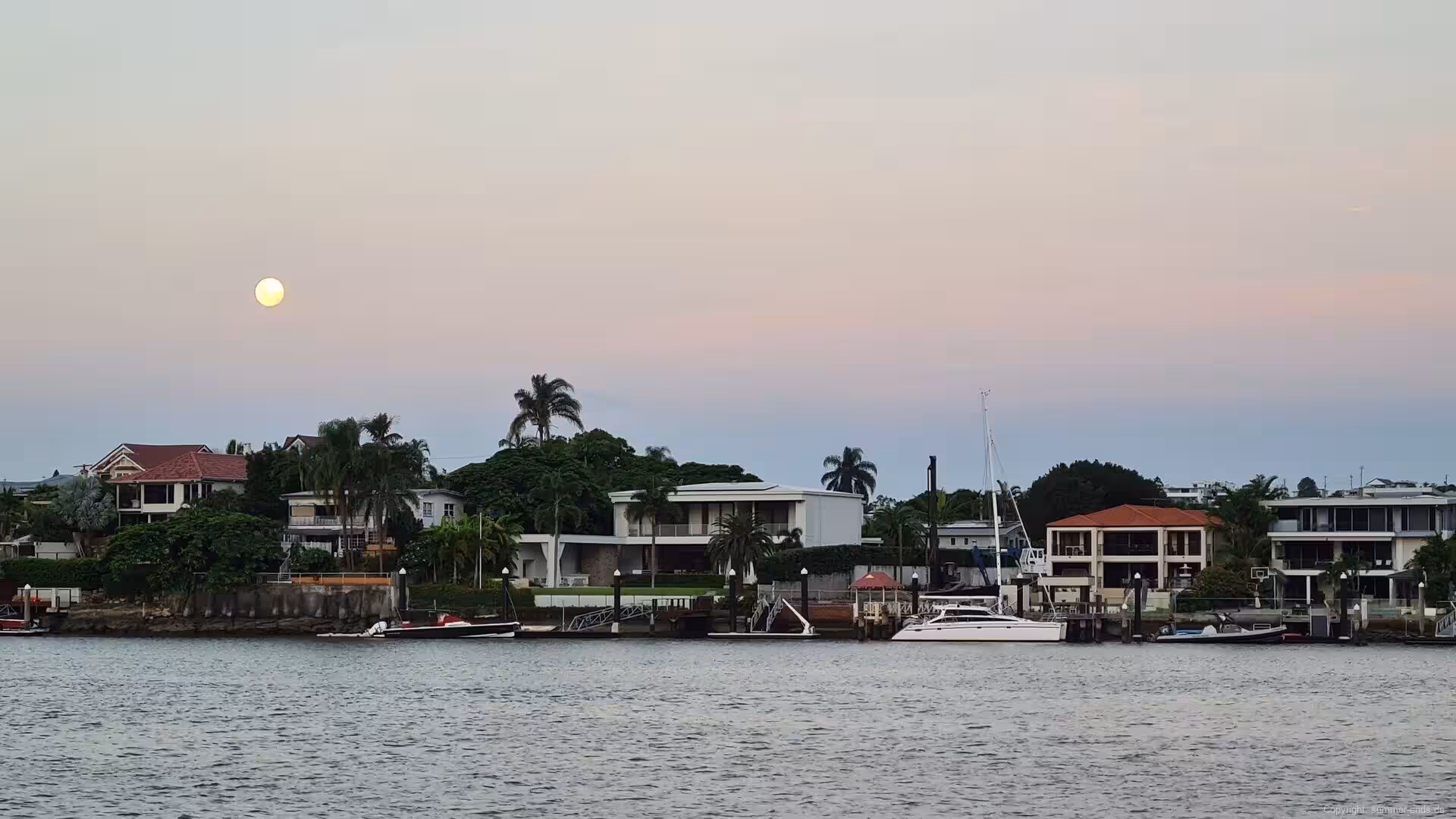 A pale moon glows over waterfront houses as dusk settles.