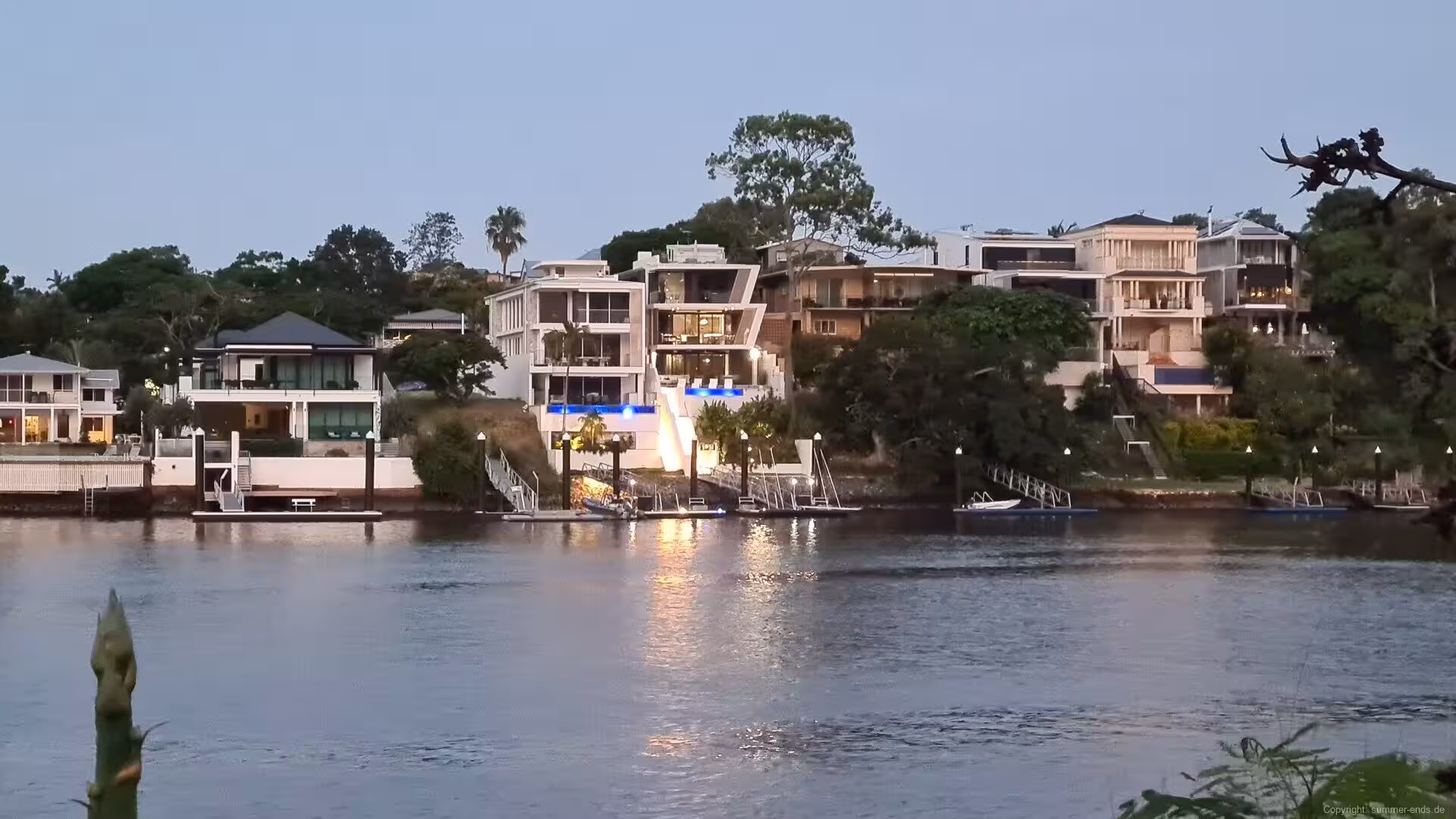 Warm lights begin to glow among waterside houses at dusk.