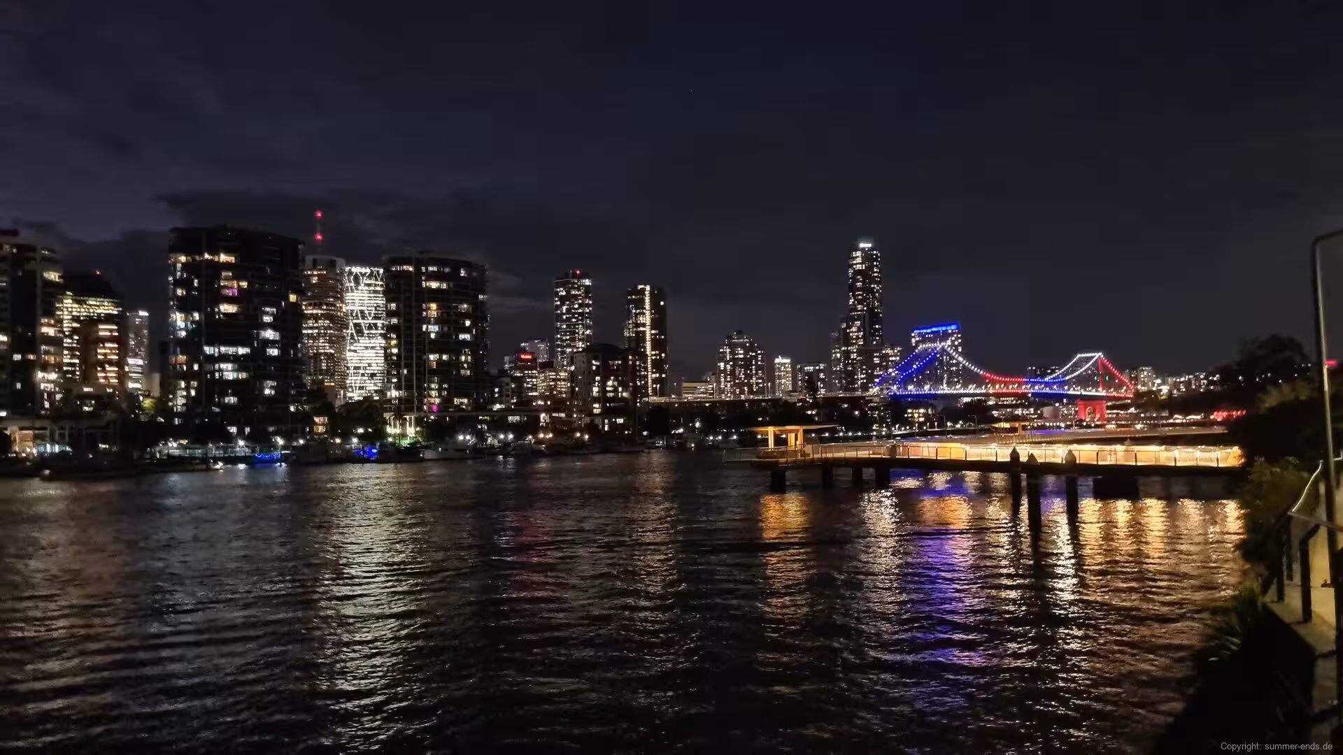 A steel bridge glows in blue and red across a night river beneath lit towers.