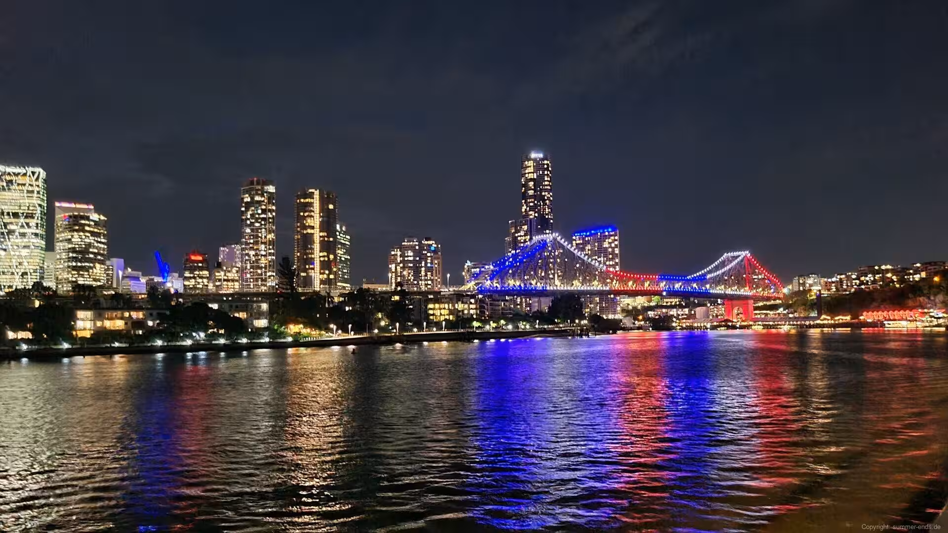 Blue and red bridge lights shimmer across a dark river in front of city towers.