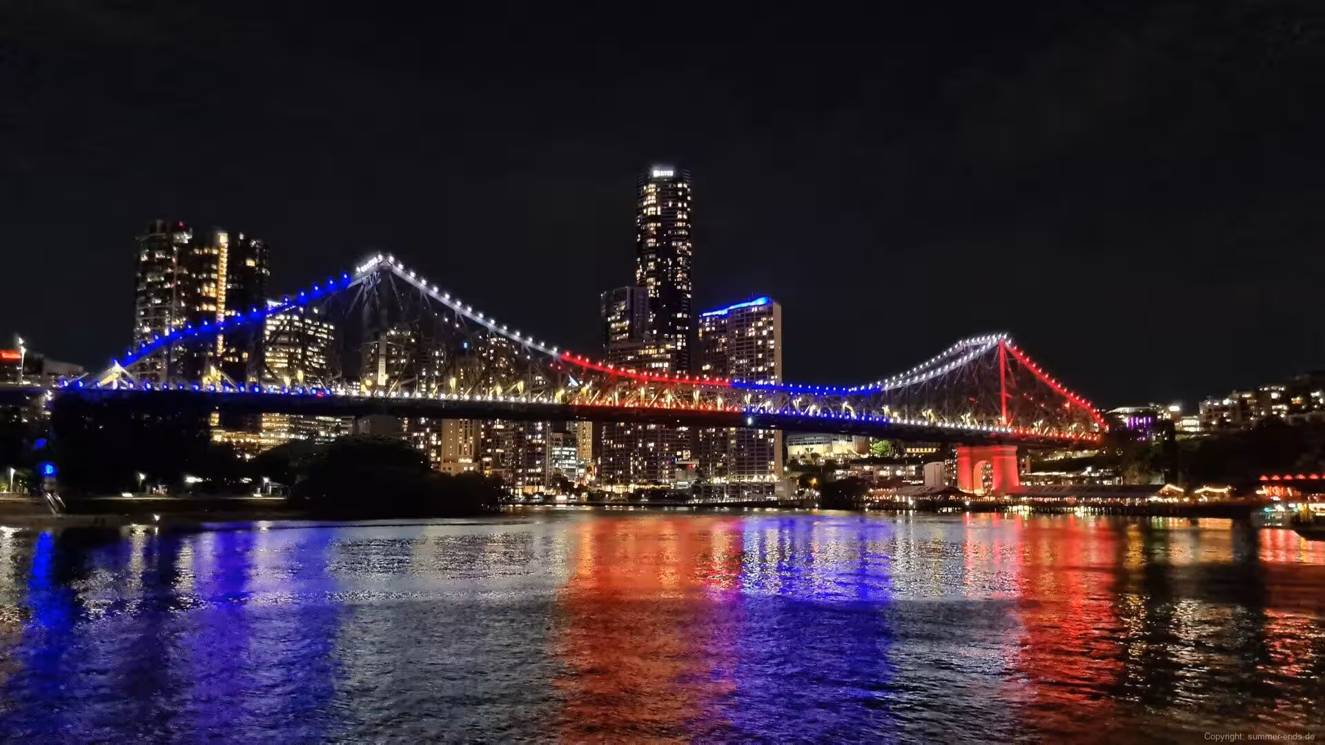 An illuminated steel bridge with blue and red lights spans a river at night.