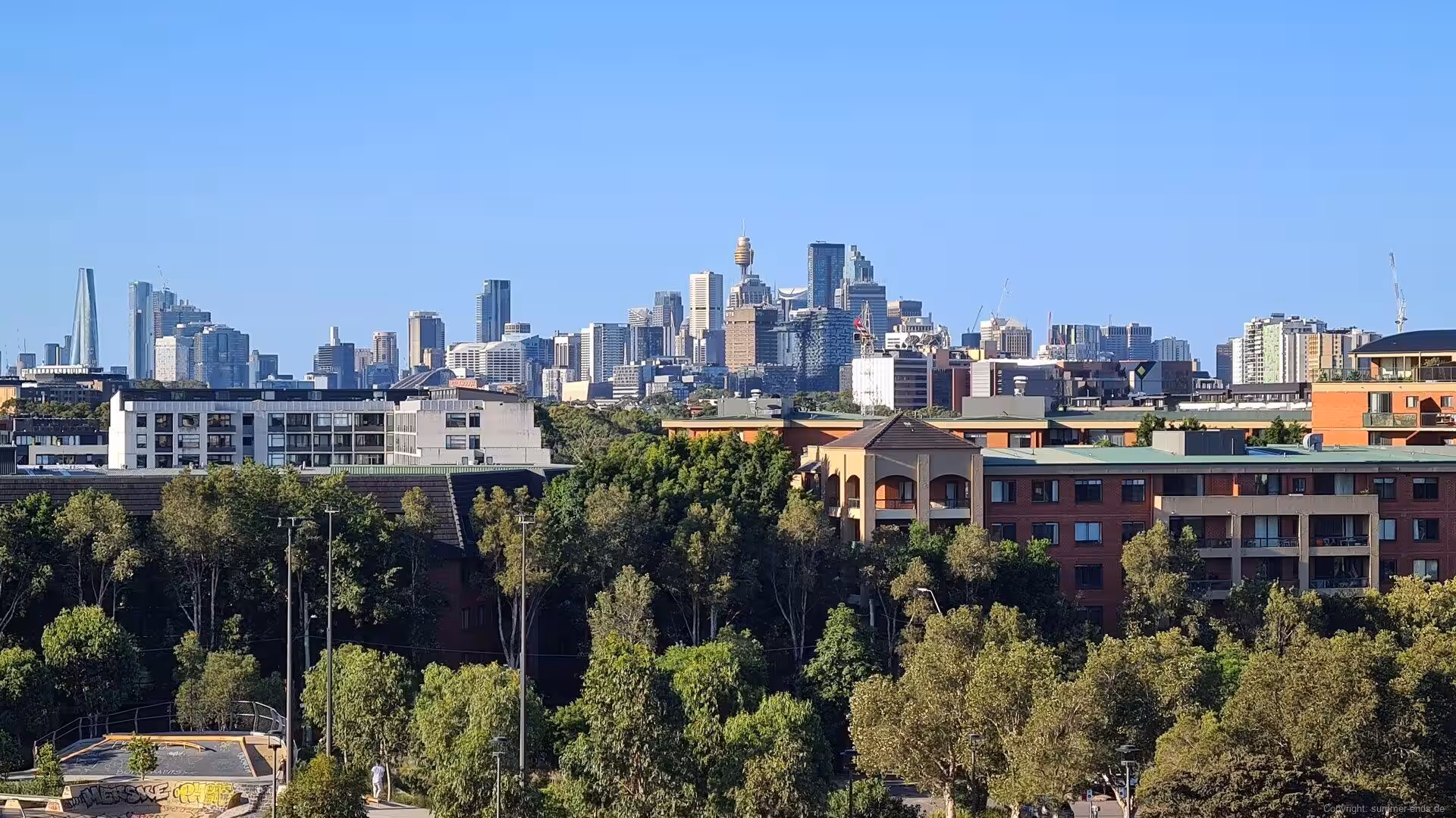 A broad view of downtown high-rises rising behind green trees and brick apartments under a clear blue sky.
