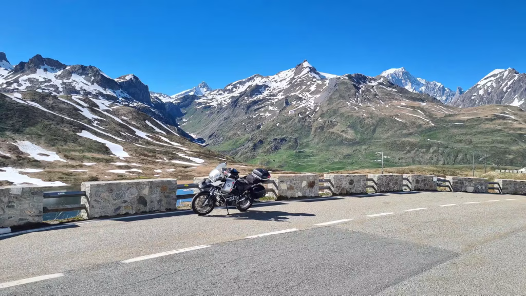 A touring motorcycle is parked by a low stone wall on a mountain pass, with snow-streaked peaks behind.