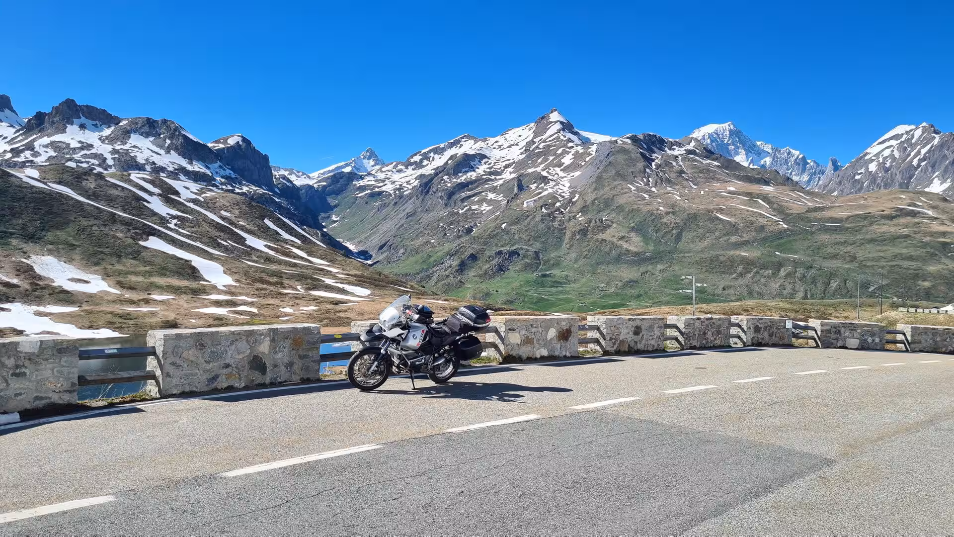 A touring motorcycle is parked by a low stone wall on a mountain pass, with snow-streaked peaks behind.