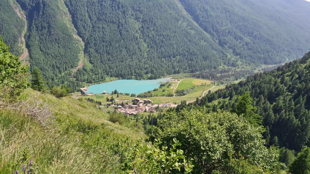 Looking down a forested slope to a bright turquoise alpine lake, with a cluster of stone houses at its shore surrounded by dense conifer woods.