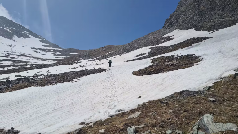 A hiker with a backpack walks up a sloping snowfield at a mountain pass, leaving a trail of footprints between rocky, snow-streaked slopes.