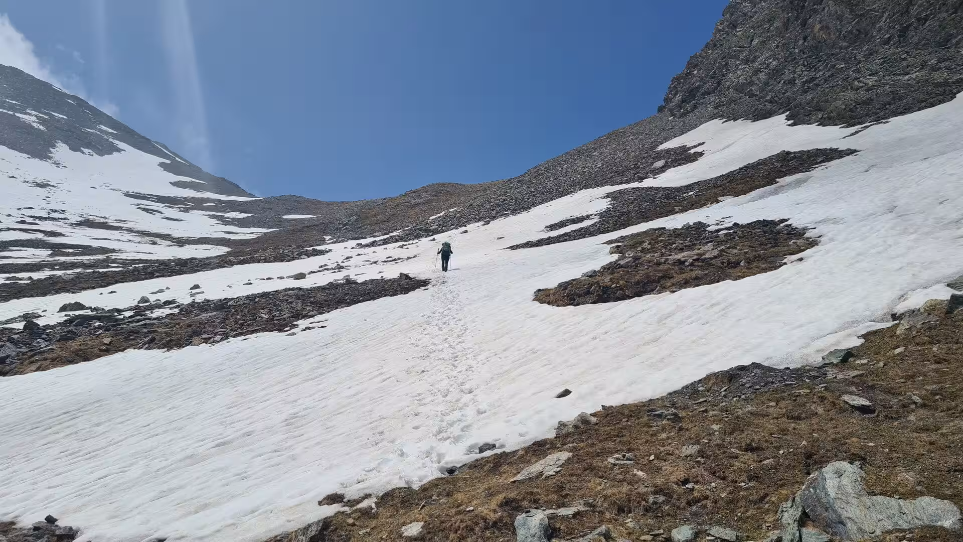A hiker with a backpack walks up a sloping snowfield at a mountain pass, leaving a trail of footprints between rocky, snow-streaked slopes.