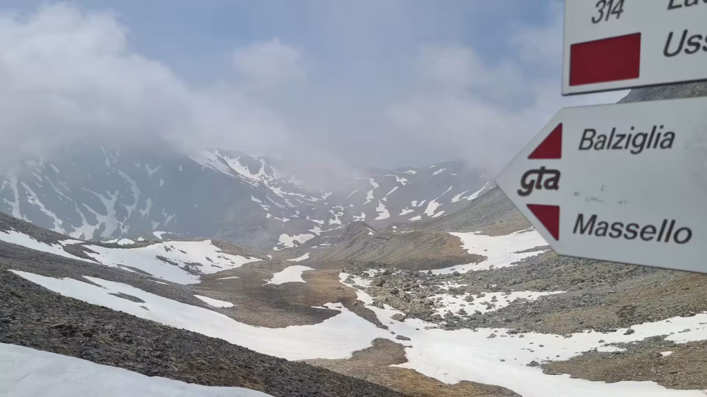 A red-and-white GTA trail signpost points toward Balziglia and Massello, standing before a snow-patched high mountain valley under low clouds.