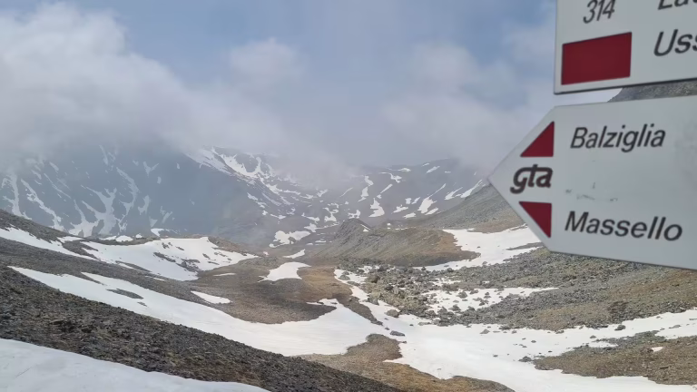 A red-and-white GTA trail signpost points toward Balziglia and Massello, standing before a snow-patched high mountain valley under low clouds.