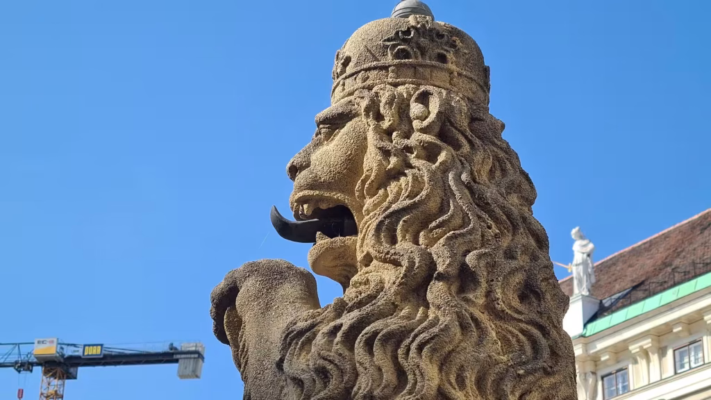 Close-up of a carved stone lion head wearing a crown, set against a bright blue sky in Vienna.