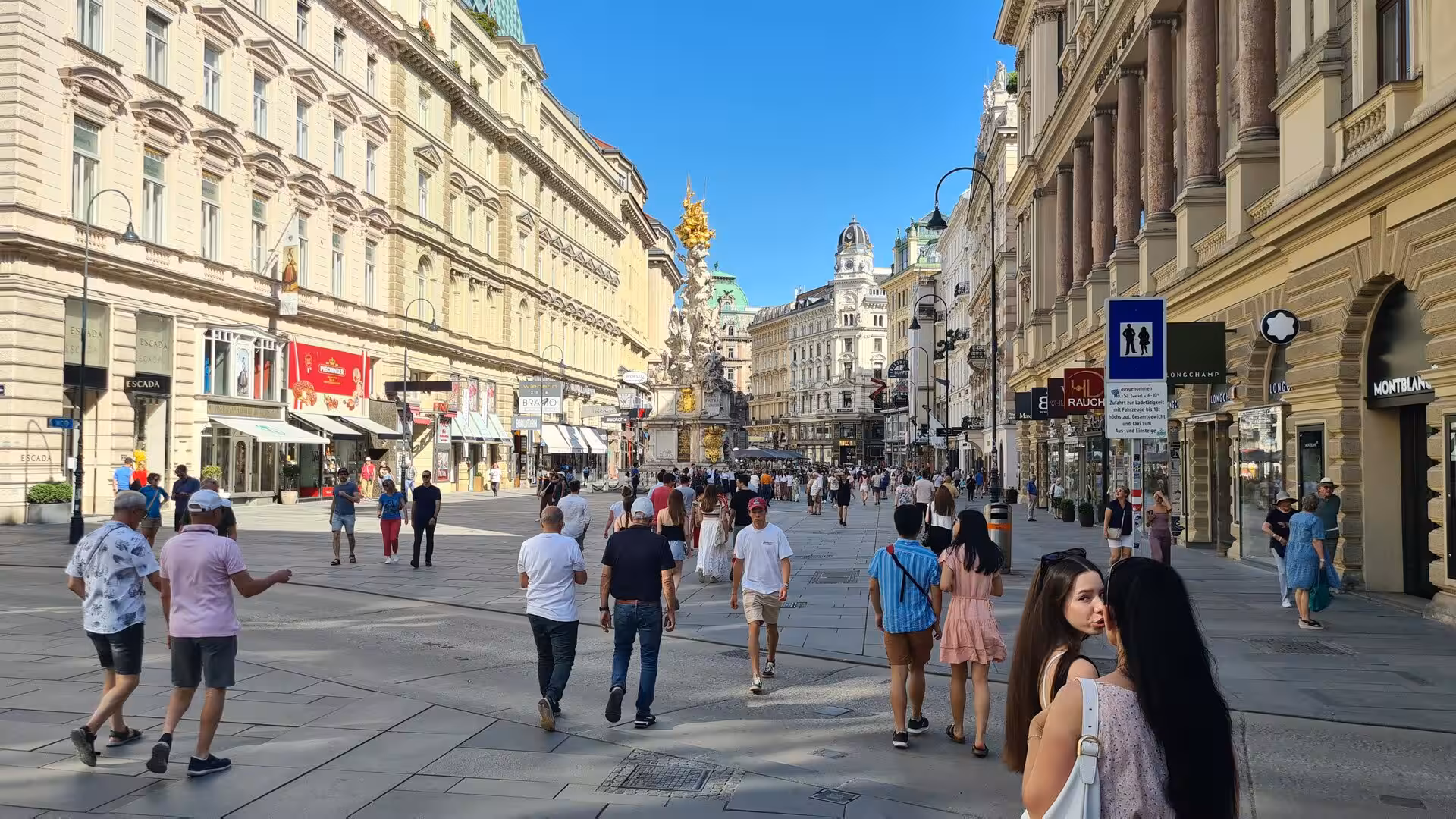 An ornate gilded monument column rises above a busy Vienna shopping street crowded with pedestrians.