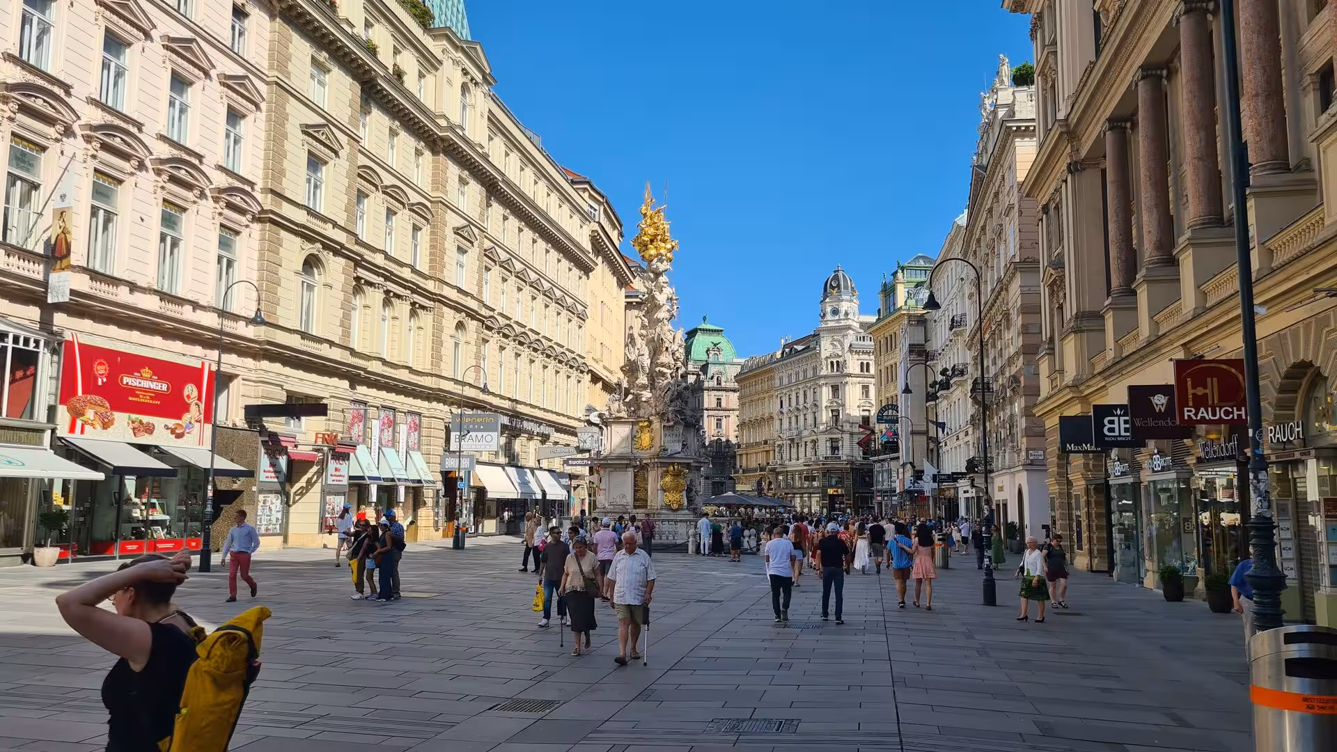 An ornamental gilded column towers over a crowded Vienna shopping street framed by historic facades.