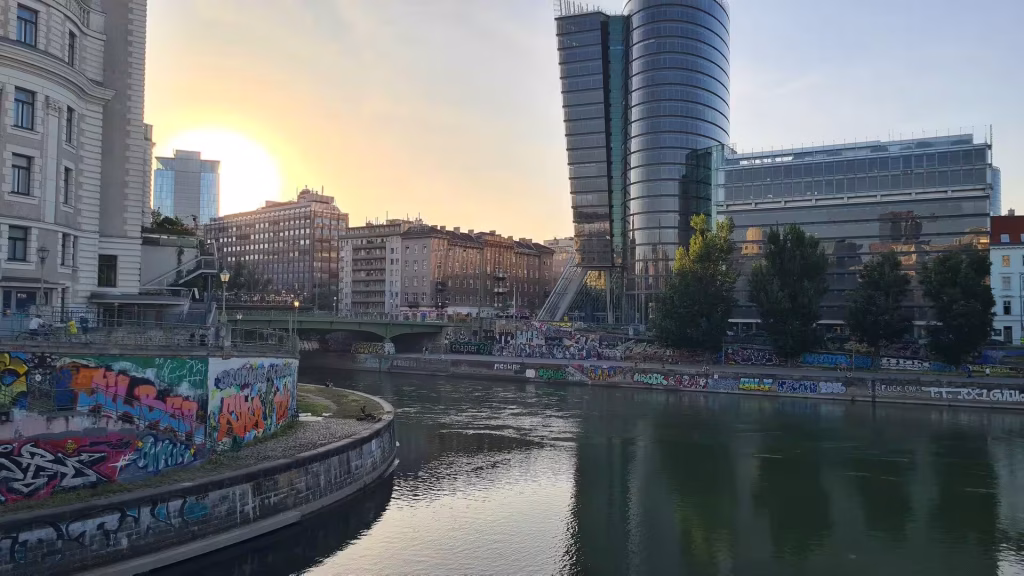 A green bridge crosses a Vienna canal with graffiti-covered walls, set against a modern glass tower at sunset.