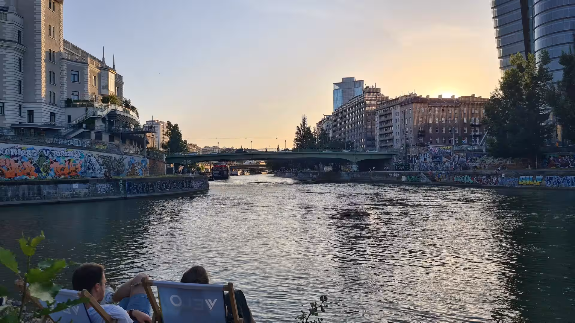 Two people lounge in chairs by a Vienna canal, with a green bridge and sunset sky beyond.