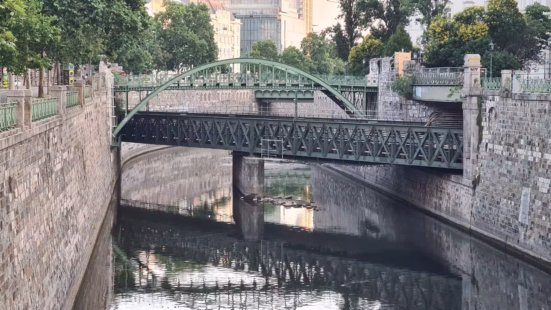 A green iron arch bridge and a lattice bridge cross a Vienna canal between stone embankments.