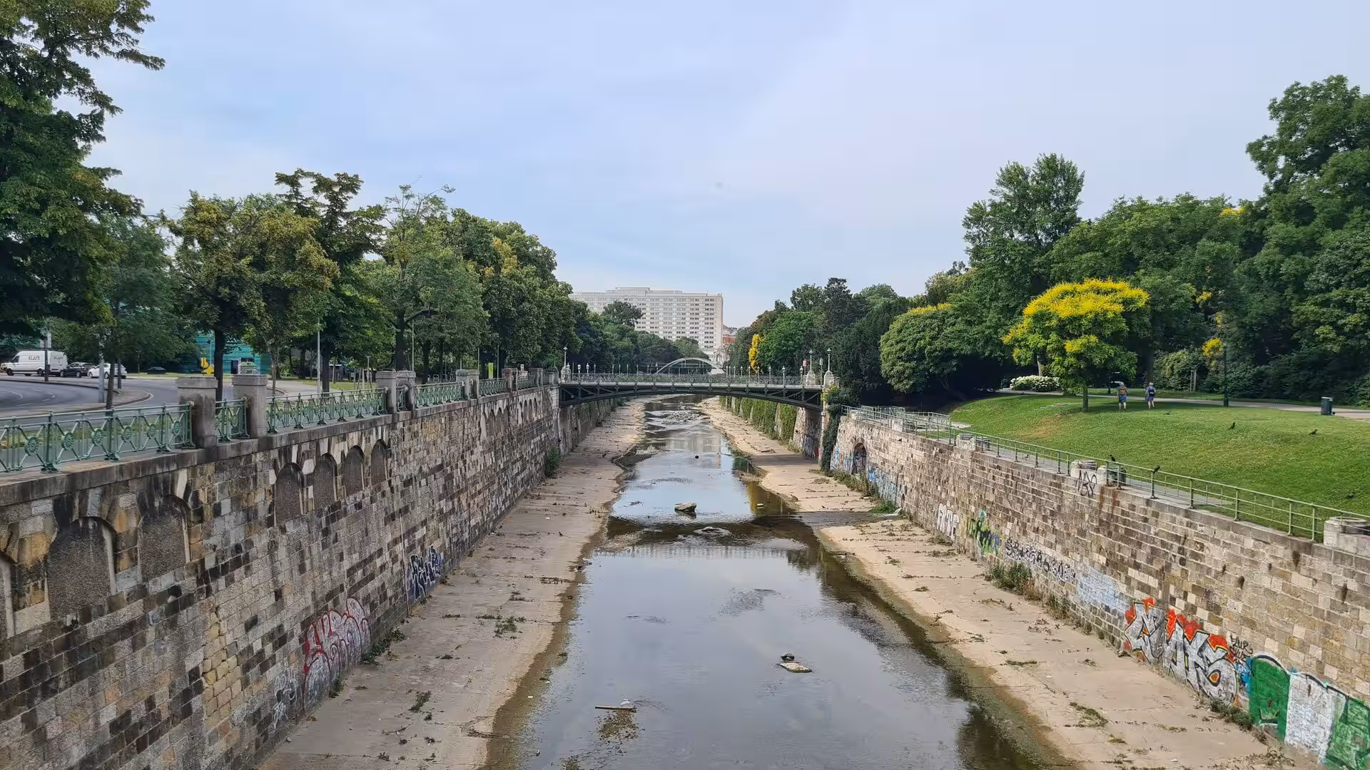 A low-water Vienna canal runs between stone embankments with graffiti and lush green banks to a distant bridge.