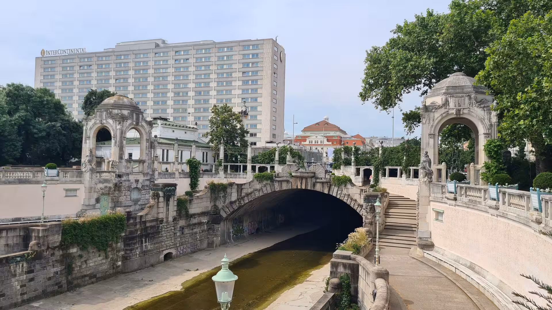An ornamental stone bridge with arched pavilions spans a Vienna waterway beneath a large hotel block.