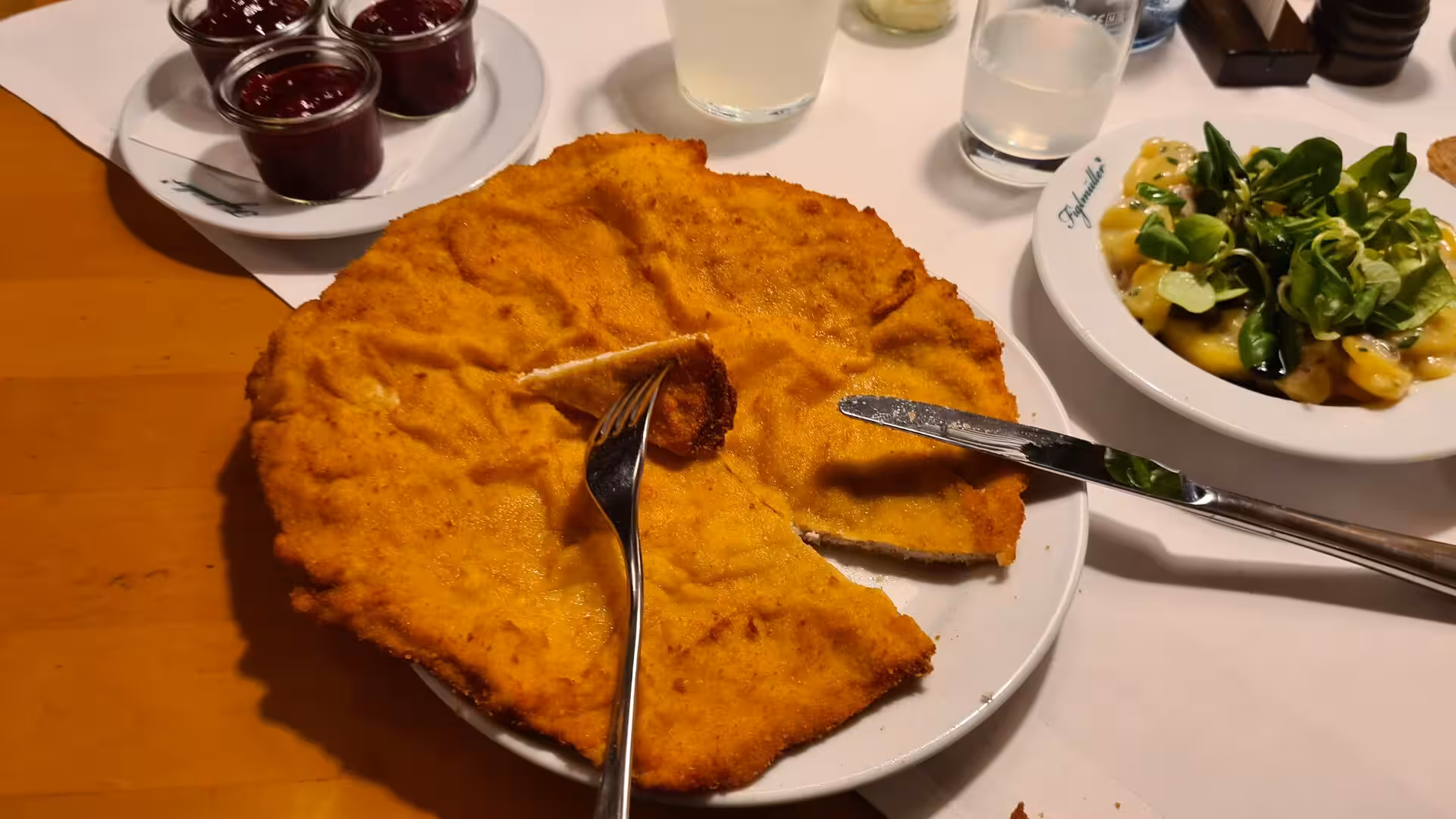 A crispy golden fried schnitzel is cut with a fork and knife, served with potato salad and small pots of dark jam on a wooden cafe table in Vienna.