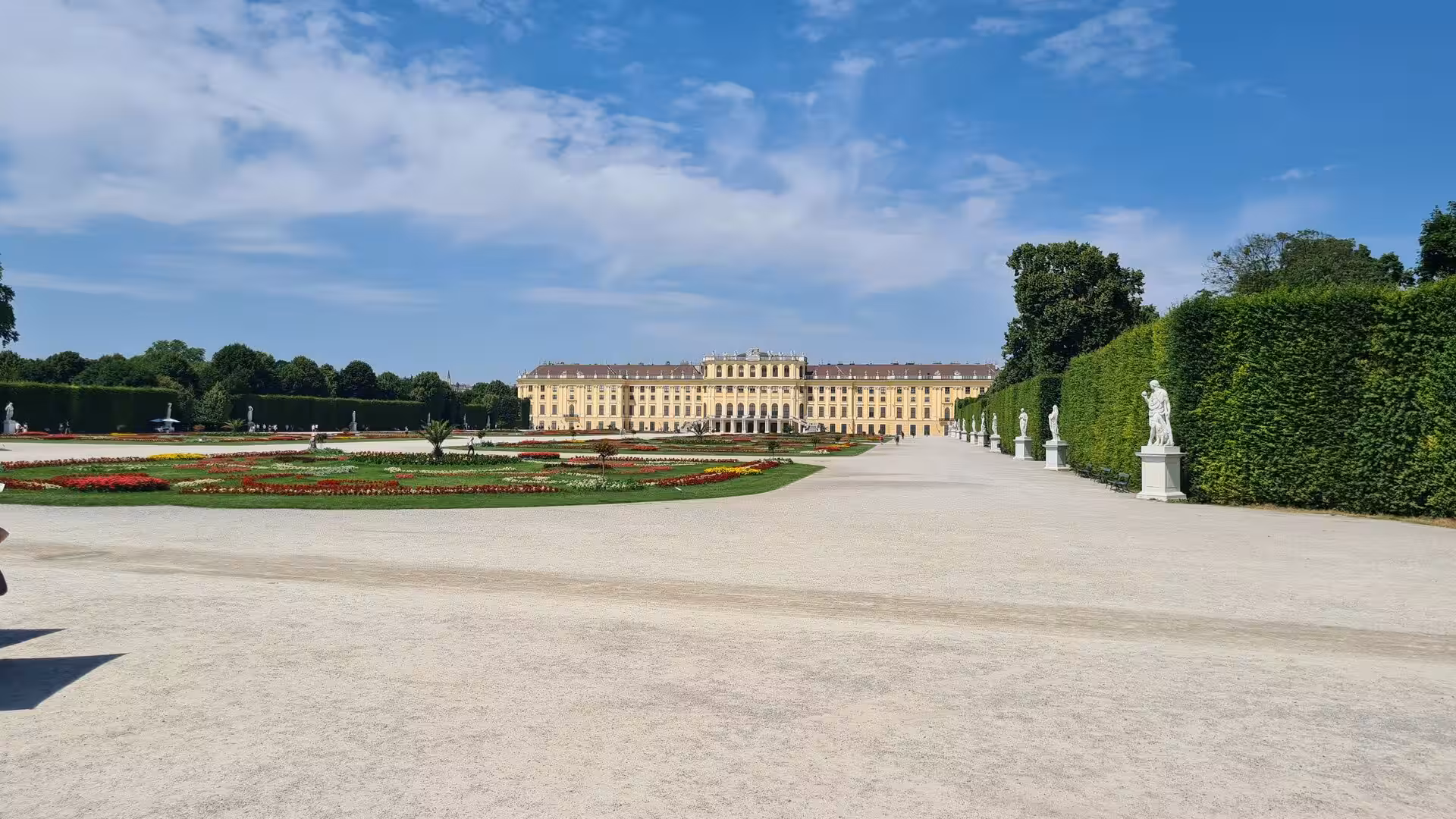 The long yellow baroque facade of Schönbrunn Palace rises across its formal gardens, bordered by trimmed hedges, flower beds and white statues under a blue sky.