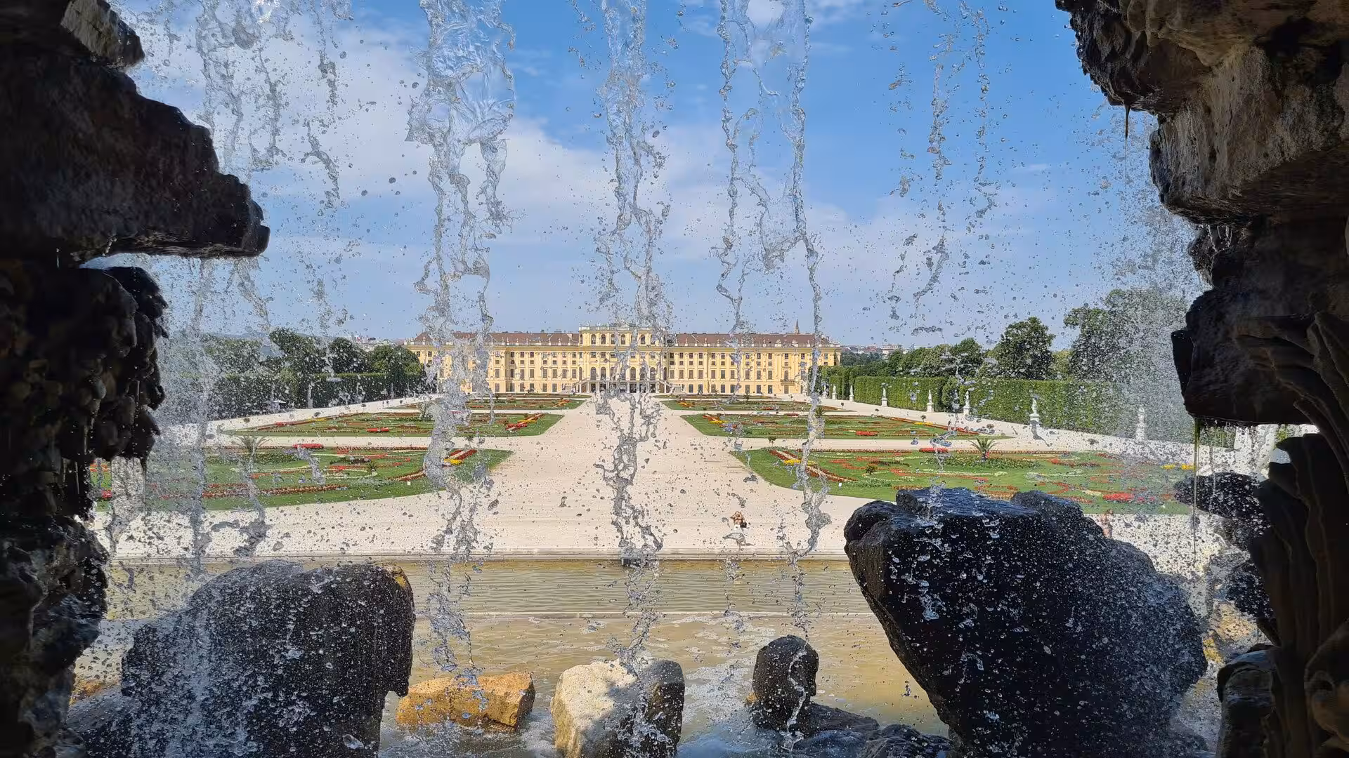 Water pours in cascading jets from a dark stone fountain, and through the gap of the splash the formal gardens and distant palace facade appear under a clear sky.