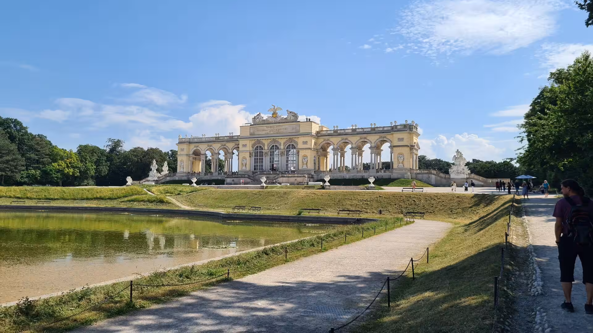 The Gloriette, a grand colonnaded building, crowns a green hill above a still pond and gravel paths dotted with visitors in warm daylight.