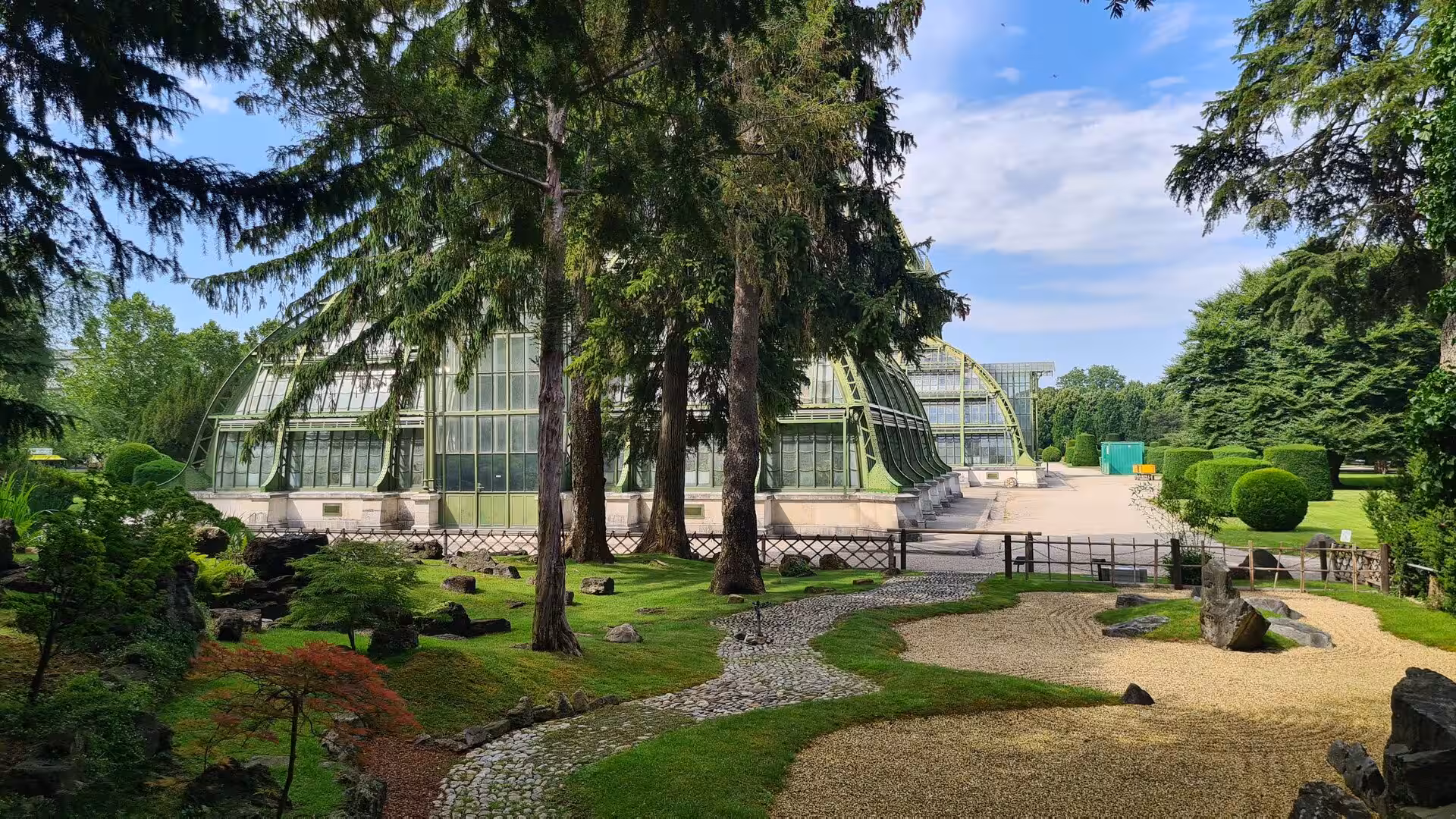 A large glass conservatory rises behind a landscaped garden with a winding gravel path, rocks and tall conifer trees beneath a blue sky.