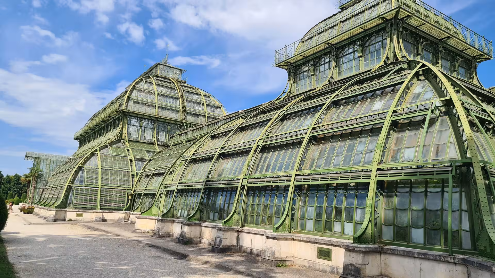 The Palm House shown close up, its pale green iron arches and tall glass panes curving gracefully toward the sky in bright daylight.