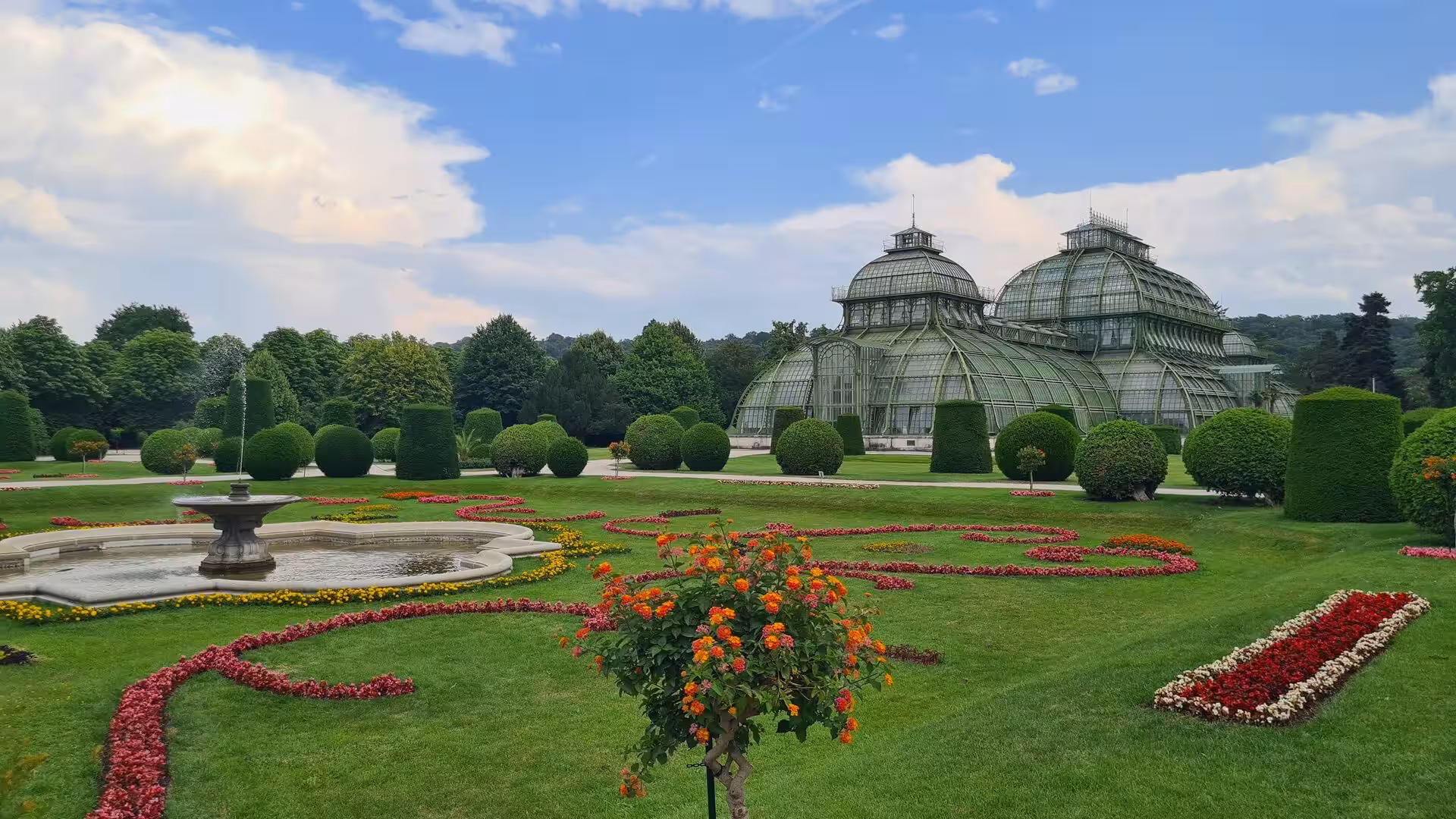Two domed glass conservatories rise beyond a formal garden with a stone fountain, clipped topiary hedges and patterned flower beds.