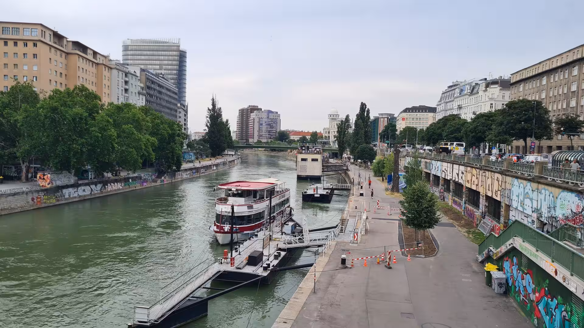 A green canal runs between graffiti-covered stone walls, a red-and-white boat moored at a pier, with a bridge and city buildings beyond.