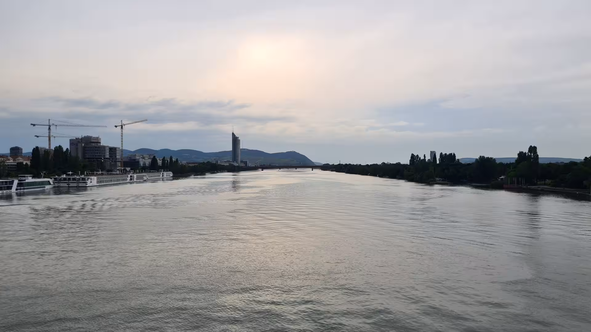 A broad calm river stretches into the distance past moored riverboats, building cranes and low green hills under a hazy, soft-lit sky.