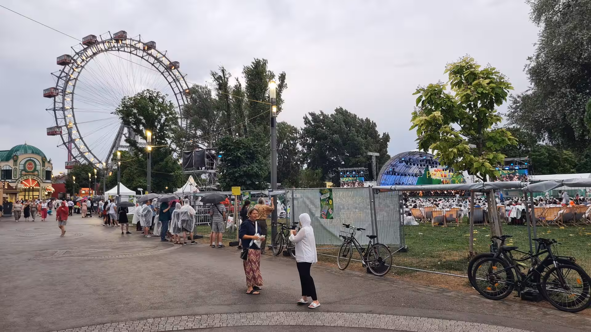Vienna's large Ferris wheel rises above a busy funfair, with a fenced-off festival area, a stage and a big crowd of visitors beneath.