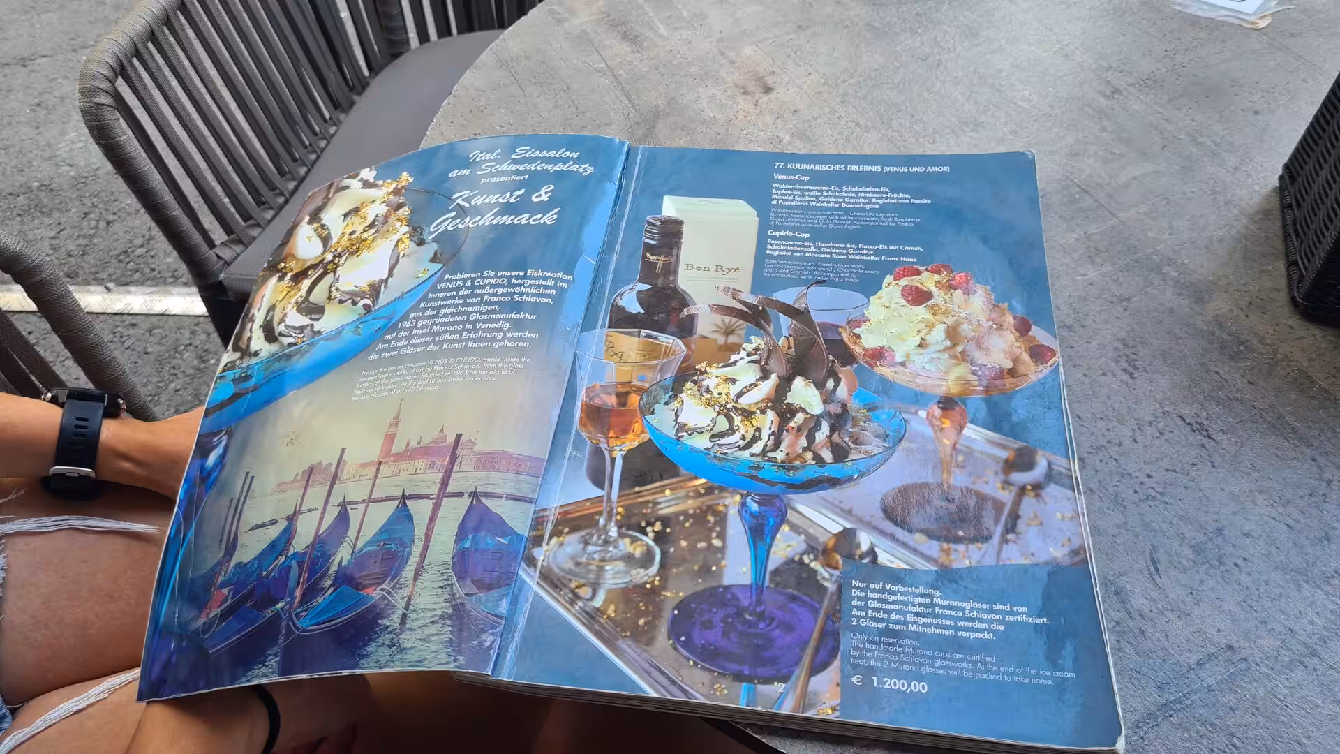An open ice cream menu showing photos of decorated sundaes and a price in euros rests on a cafe table, held by someone seated in a chair.