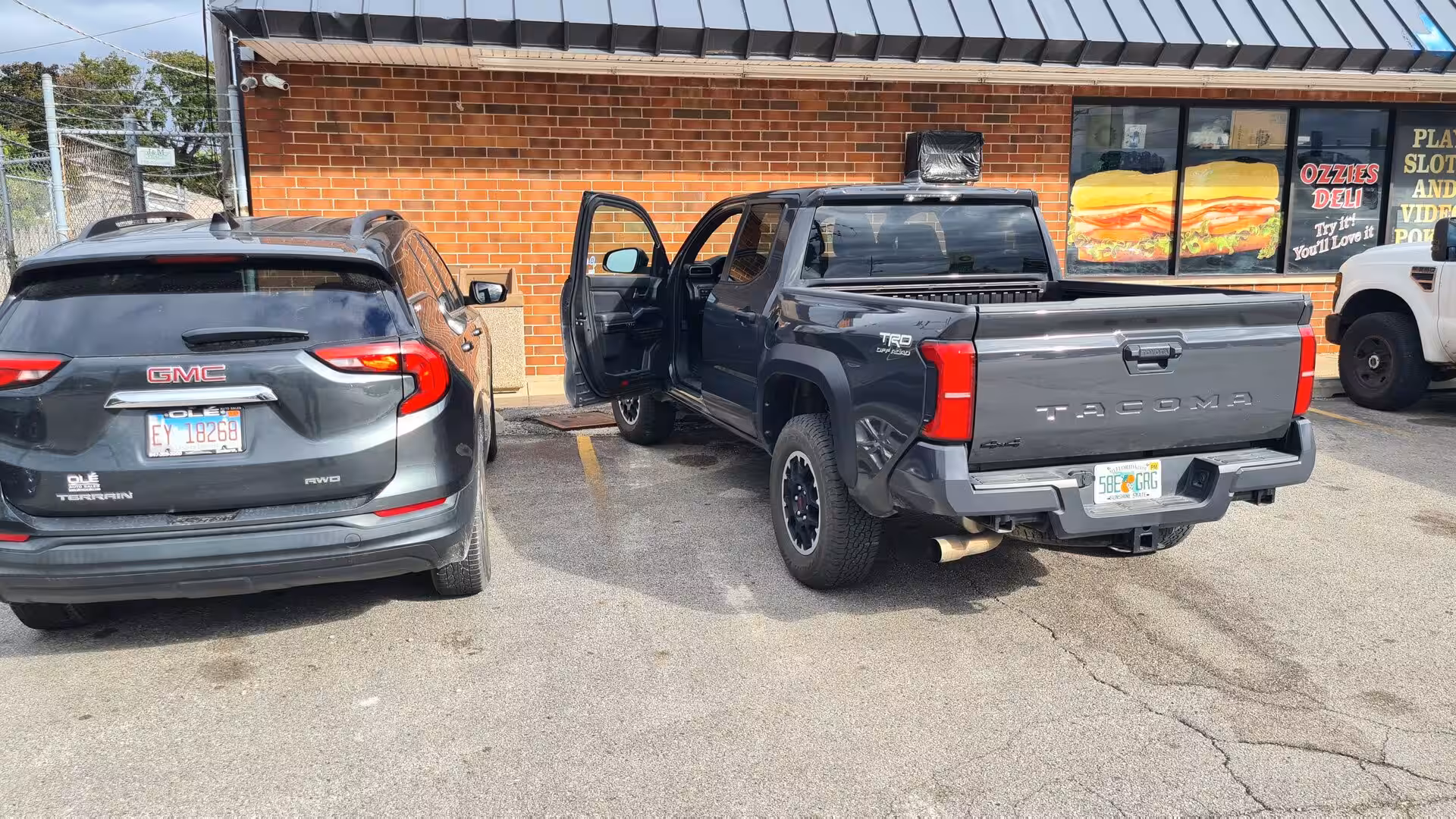 Gray Toyota Tacoma pickup and GMC Terrain SUV parked outside a roadside deli on a sunny day.
