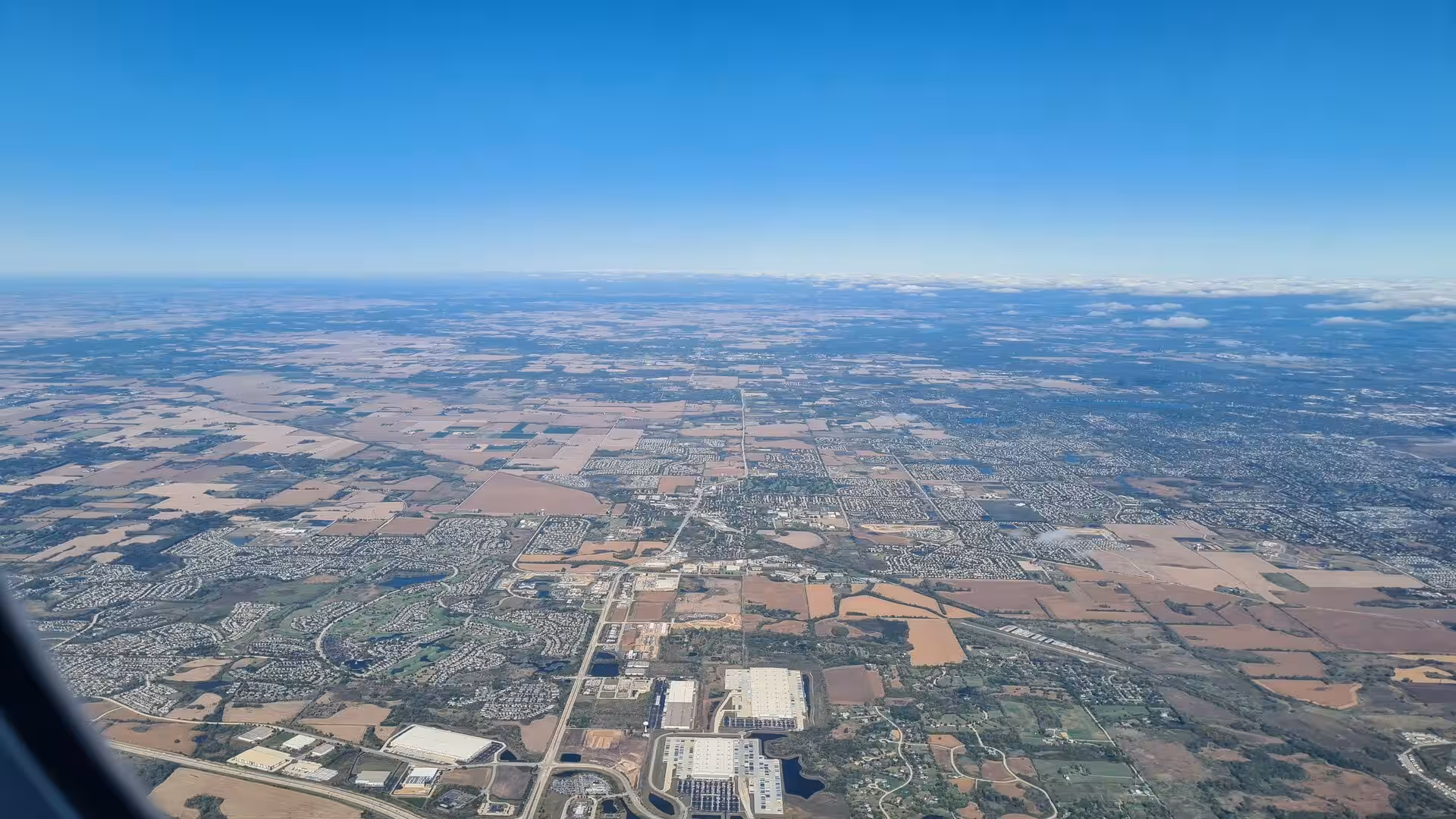 Aerial view from an airplane window showing farmland, suburban neighborhoods, roads, and industrial buildings stretching to the horizon.