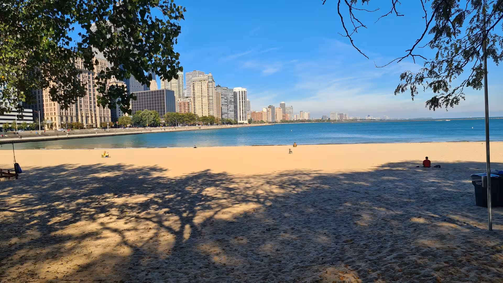 Sandy beach along Lake Michigan with Chicago’s skyline, waterfront promenade, and clear blue water on a sunny day.