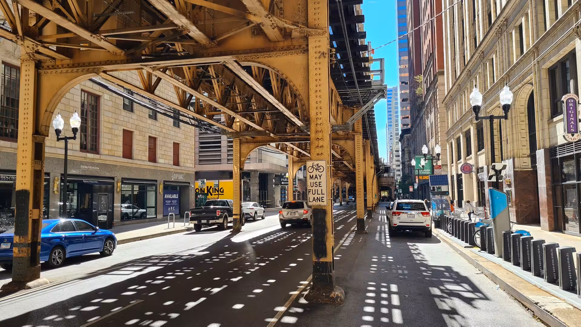 Street view beneath Chicago’s elevated CTA railway in the Loop district, with steel structures, cars, bike lanes, and surrounding historic buildings.