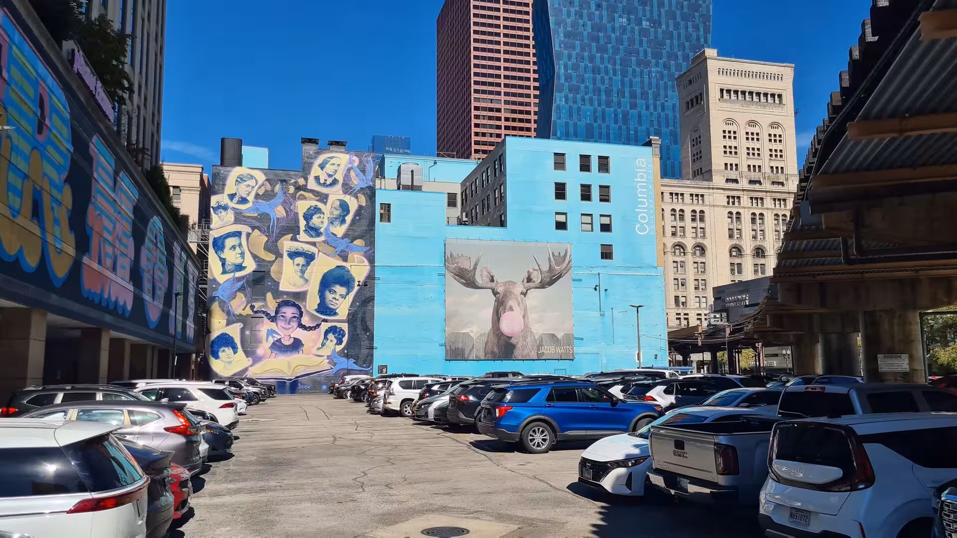 Colorful large-scale murals on building facades beside a parking area in downtown Chicago, surrounded by modern and historic high-rise architecture.