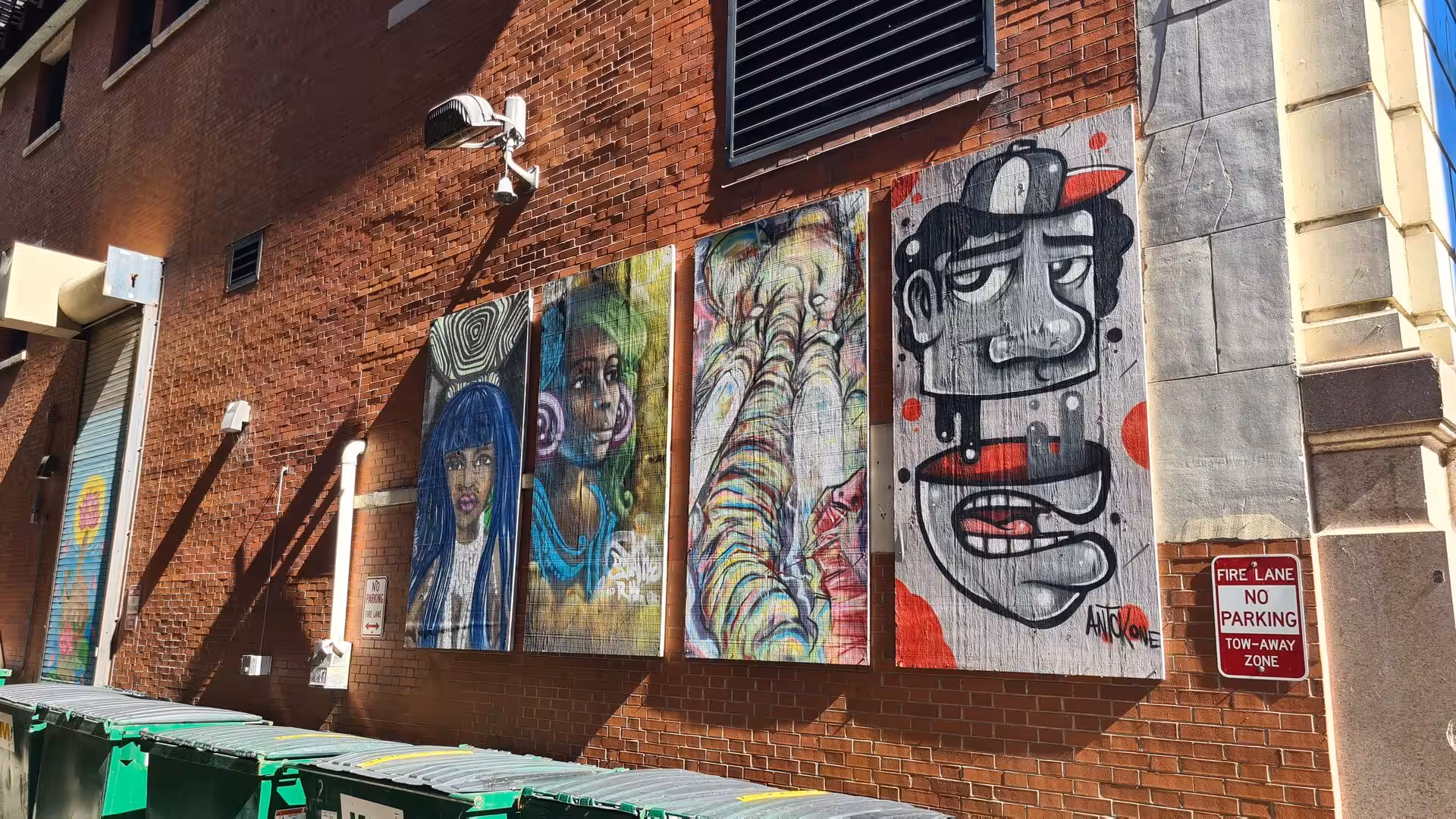 Large contemporary murals mounted on a red brick building wall in a downtown Chicago alley with urban street elements and afternoon sunlight.