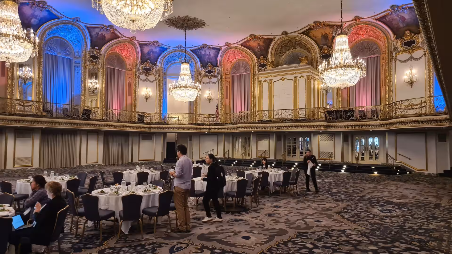 Elegant historic ballroom in Chicago featuring crystal chandeliers, ornate gold detailing, colorful uplighting, and banquet tables.