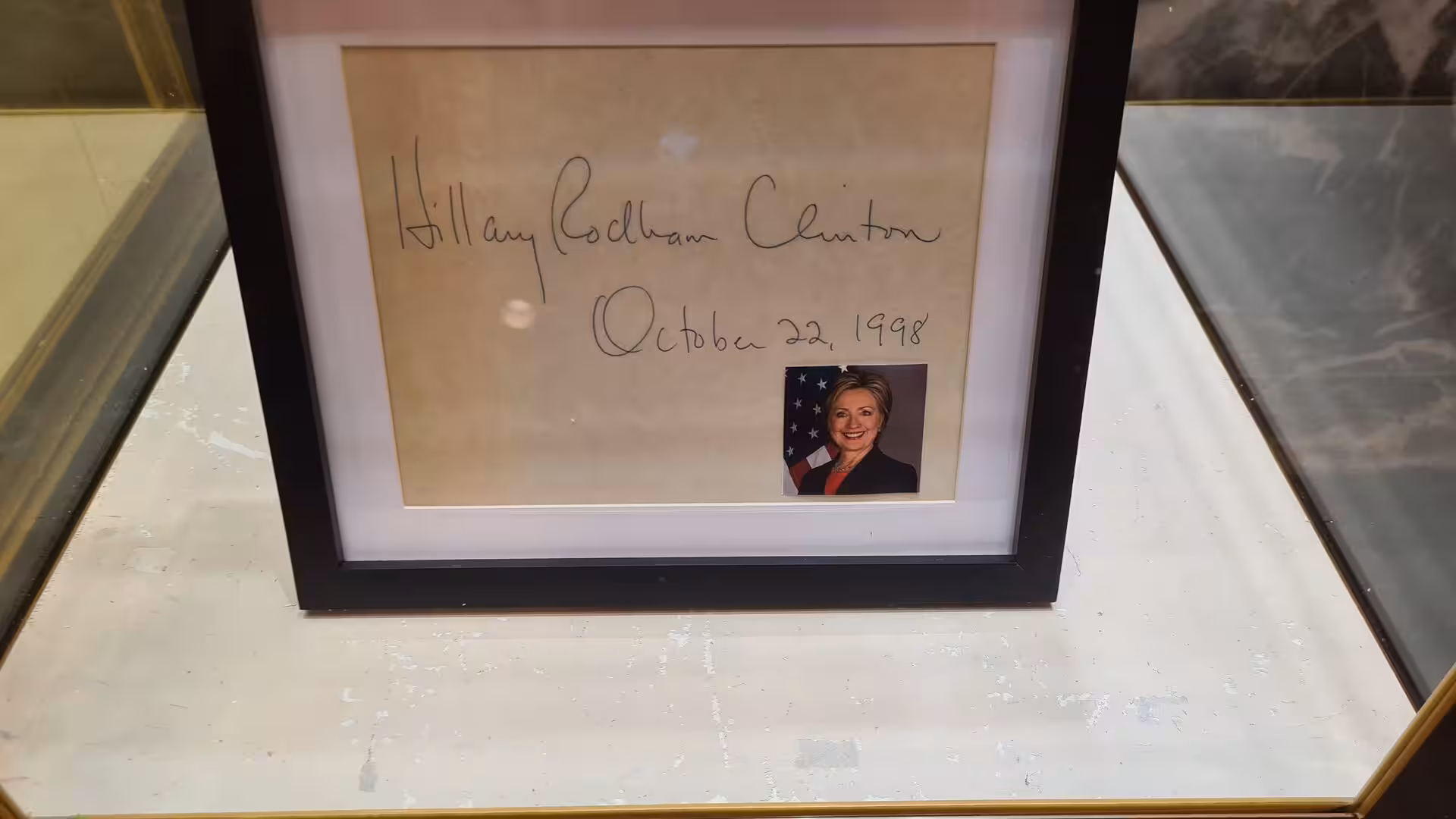 Framed handwritten signature reading “Hillary Rodham Clinton” with a dated inscription and a portrait photograph displayed inside a glass showcase.