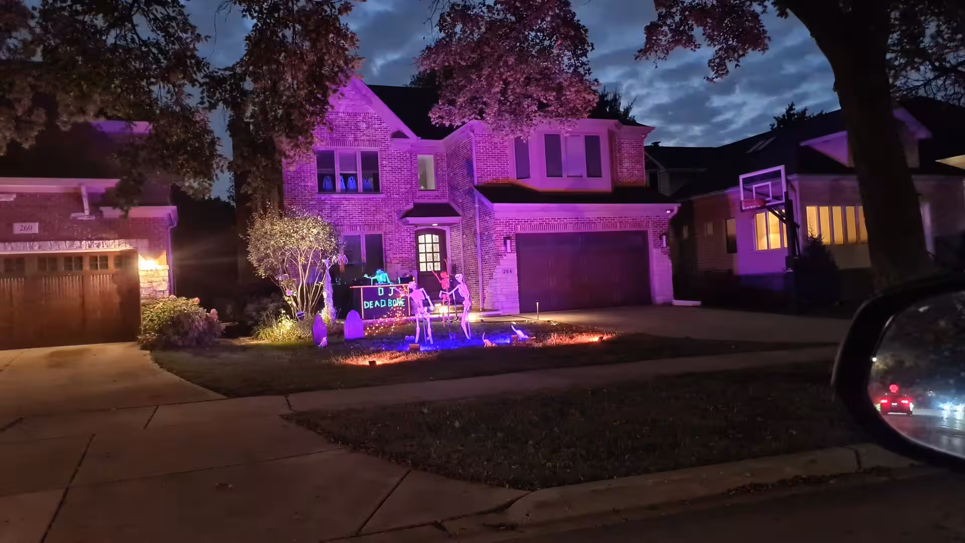 Suburban brick house illuminated with purple lighting and decorated with Halloween skeletons, glowing props, and themed front yard displays at dusk.