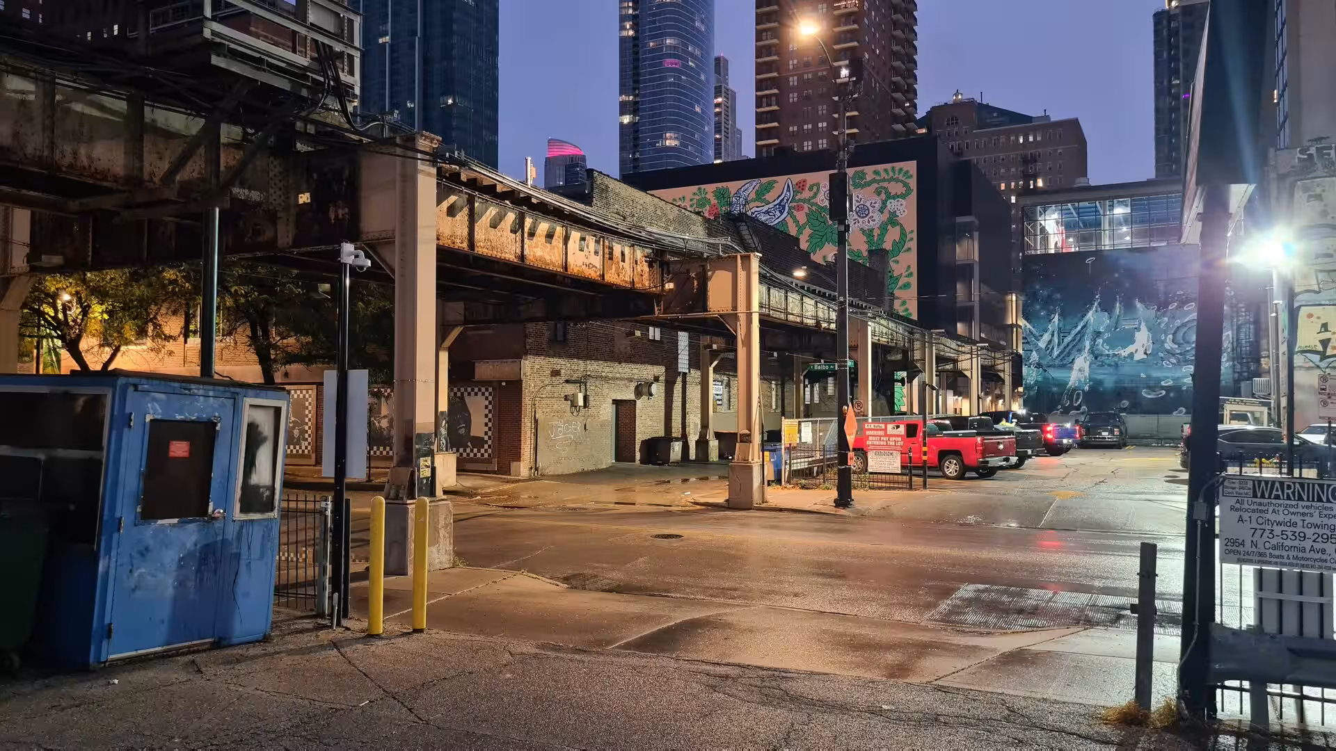 Nighttime city scene beneath Chicago’s elevated railway featuring industrial steel structures, illuminated murals, parked vehicles, and downtown skyscrapers.