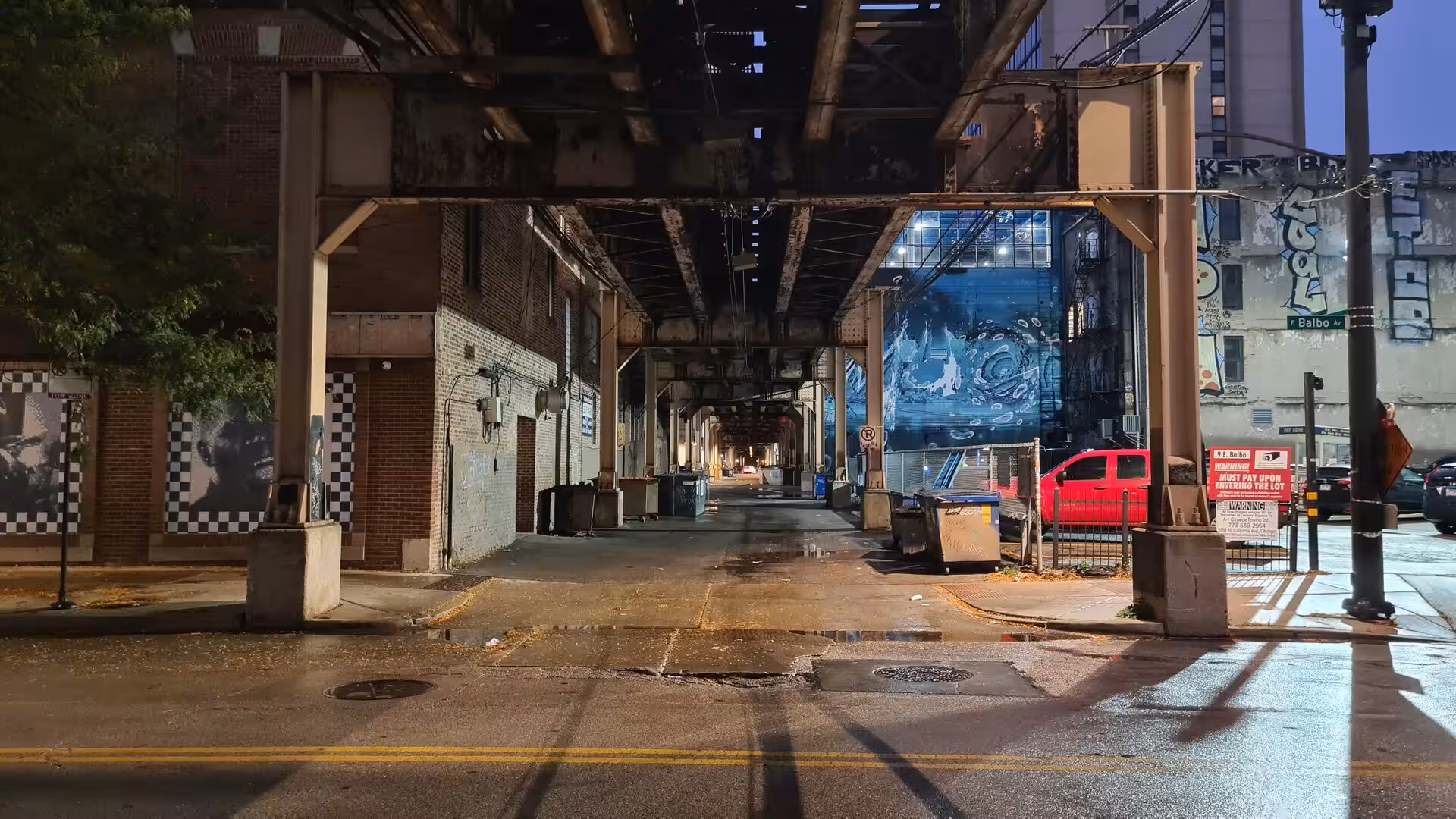 Night view beneath Chicago’s elevated railway showing steel structures, wet pavement reflections, brick buildings, urban murals, and a quiet downtown side street.