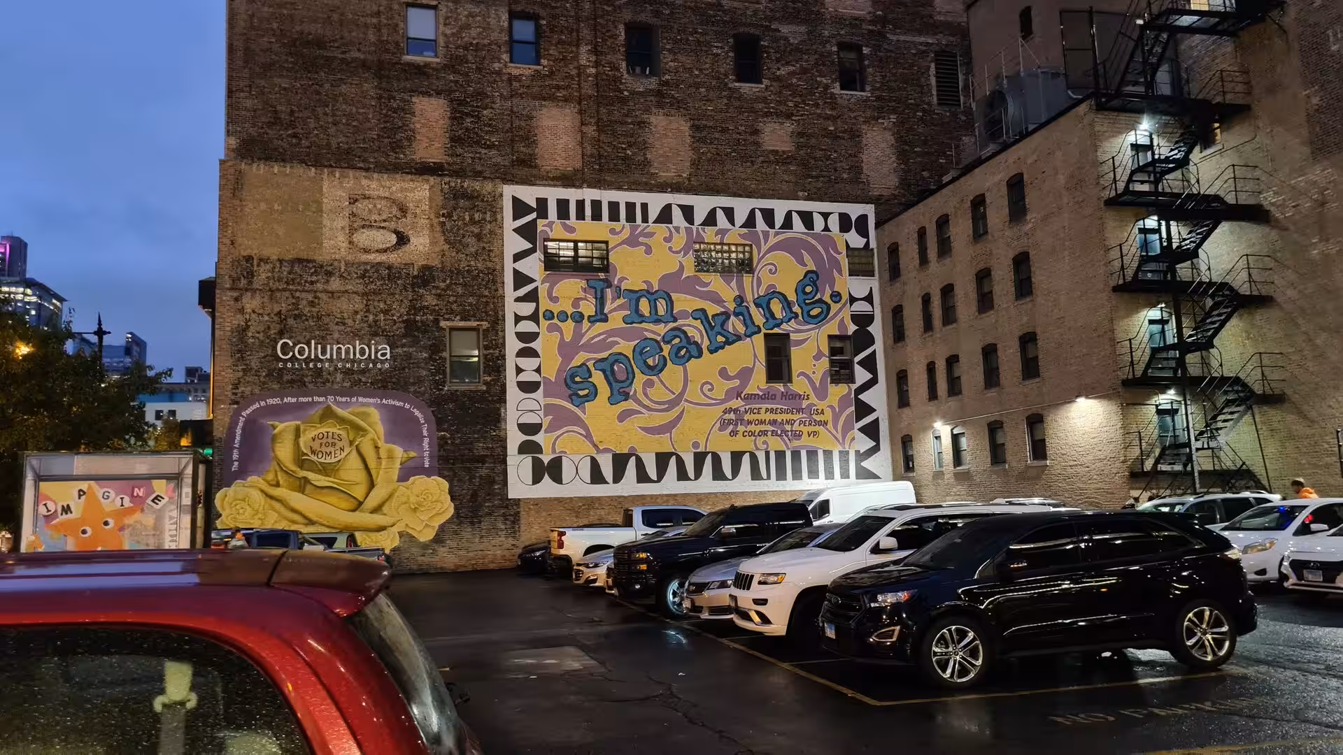 Large illuminated mural reading “I’m speaking” on a brick building beside a parking area in downtown Chicago during the evening.