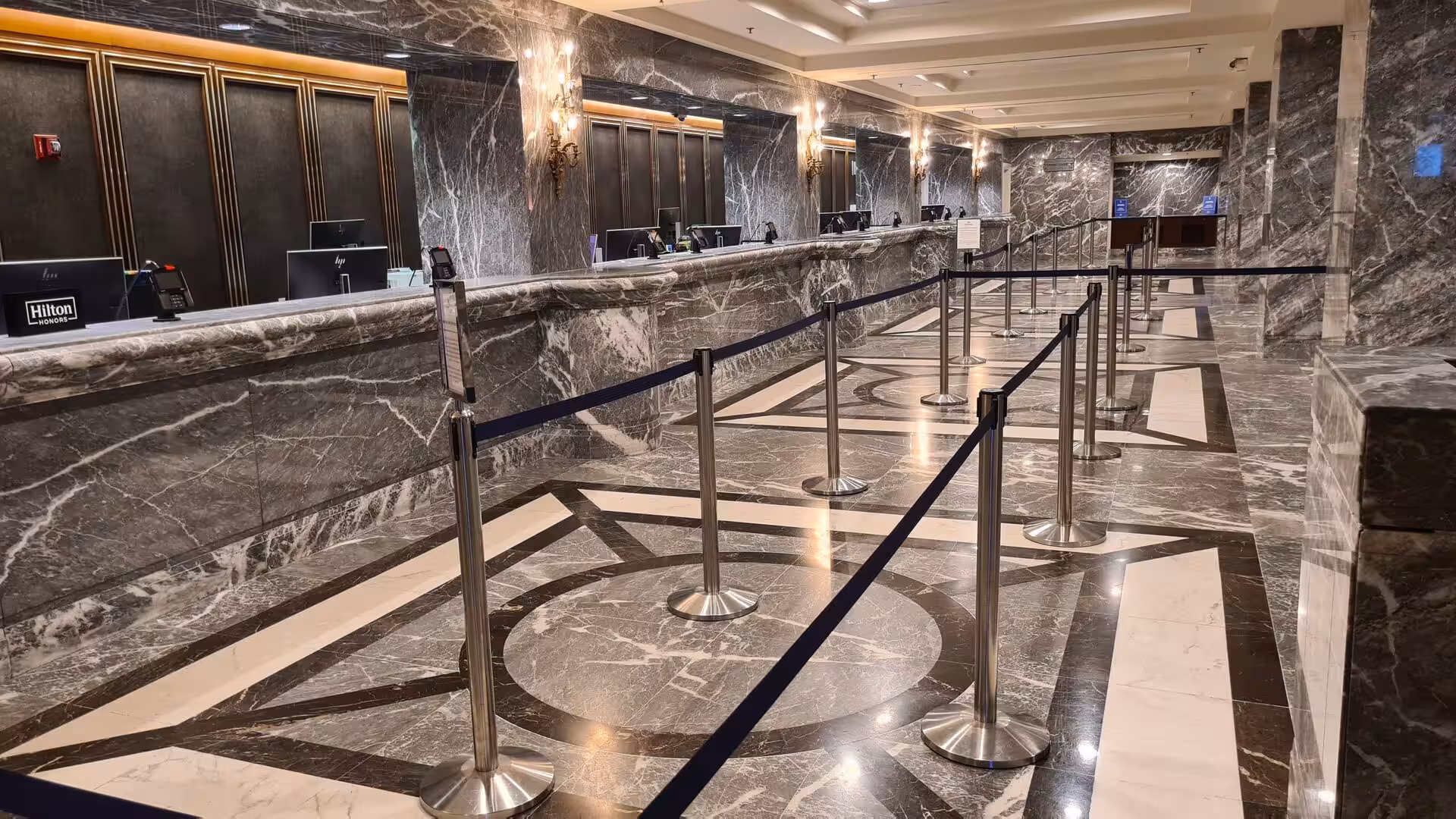 Elegant hotel reception area featuring dark marble walls, geometric marble flooring, and a long check-in counter with queue barriers.